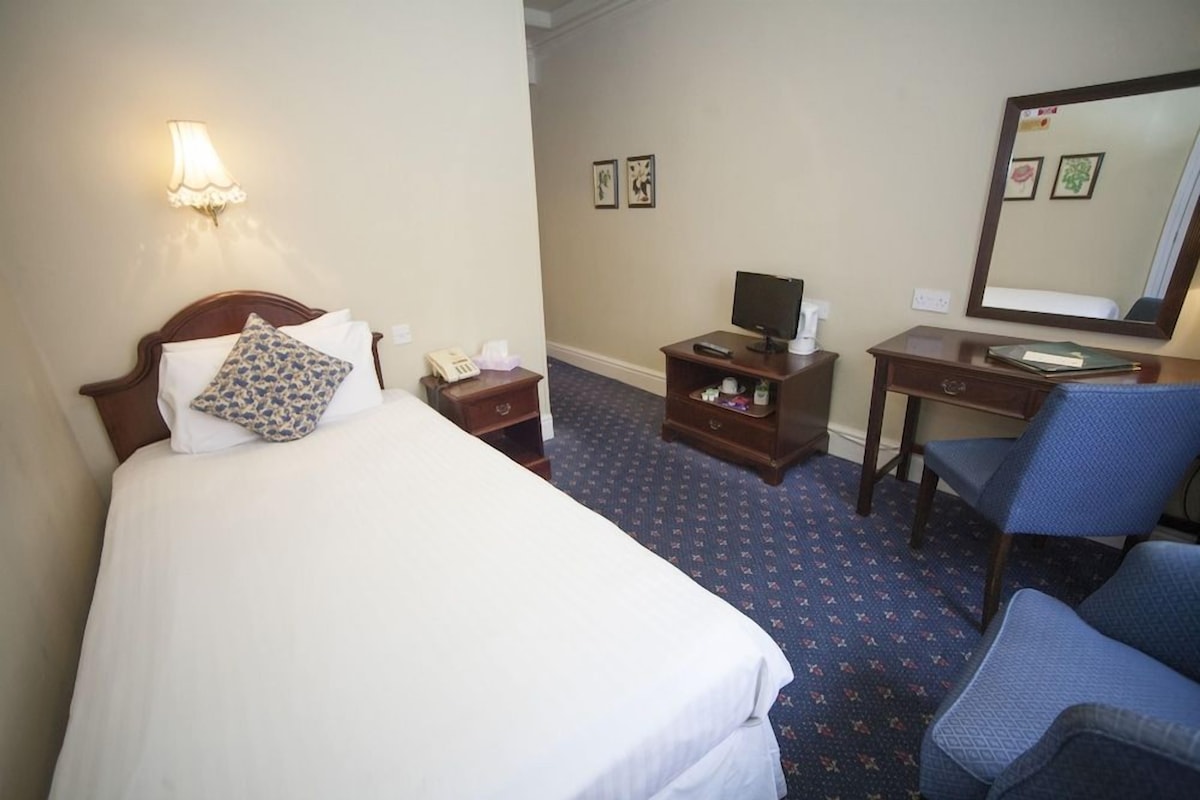 Ein Hotelzimmer mit Bett, Nachttisch, Lampe, Schreibtisch, Stuhl und einem großen Spiegel.
