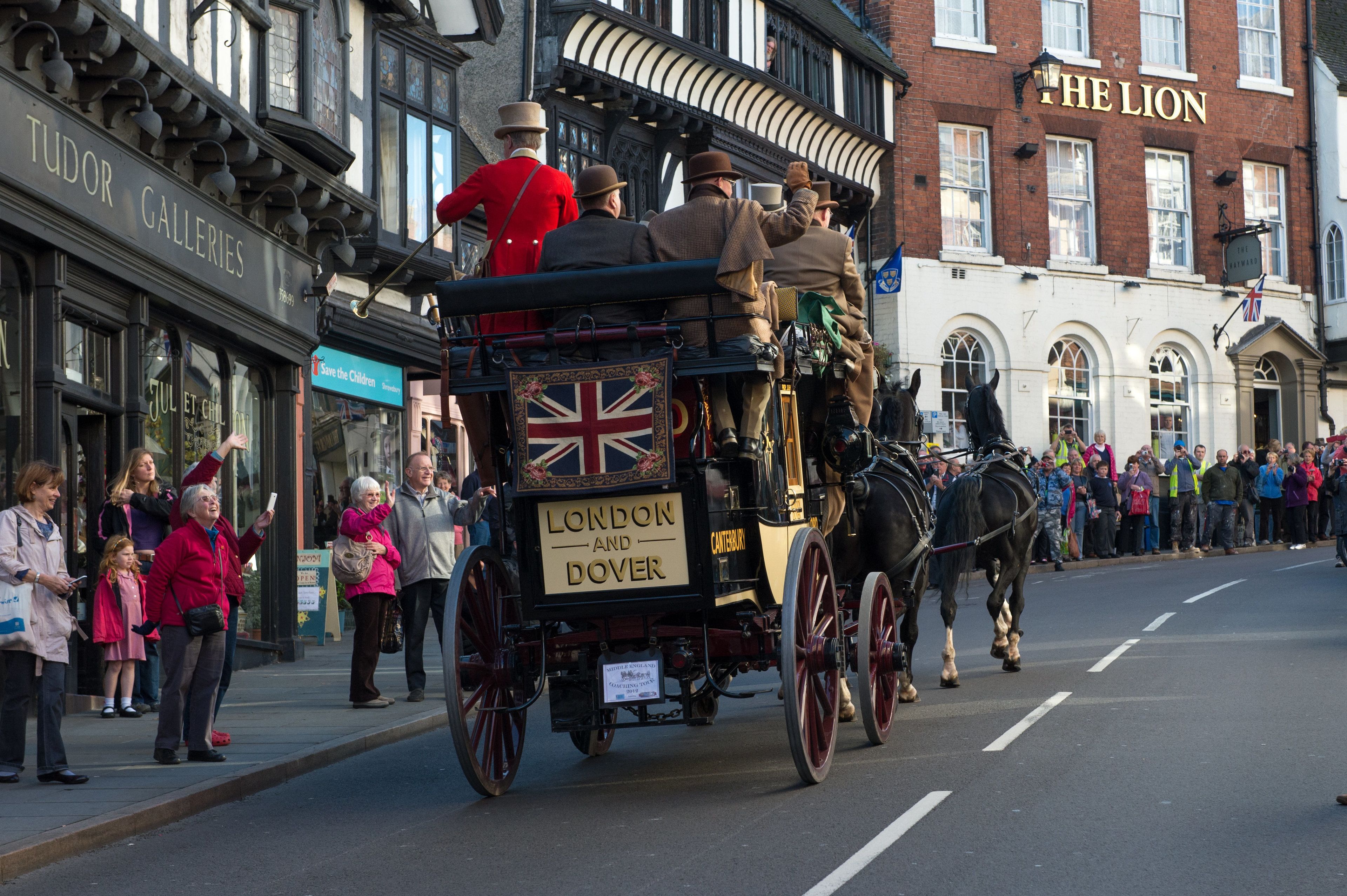 A horse-drawn carriage with passengers and a Union Jack flag, moving down a street lined with shops and buildings.