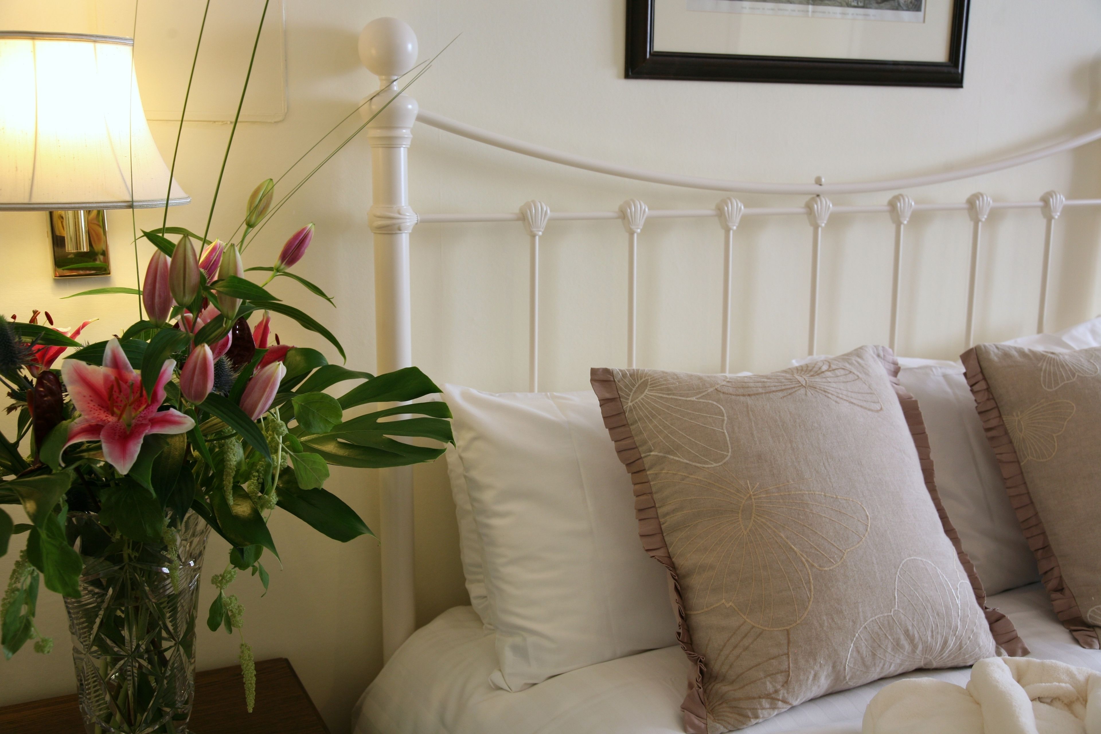 A neatly made bed with white and beige pillows, a clear vase with flowers, and a lamp on a bedside table.