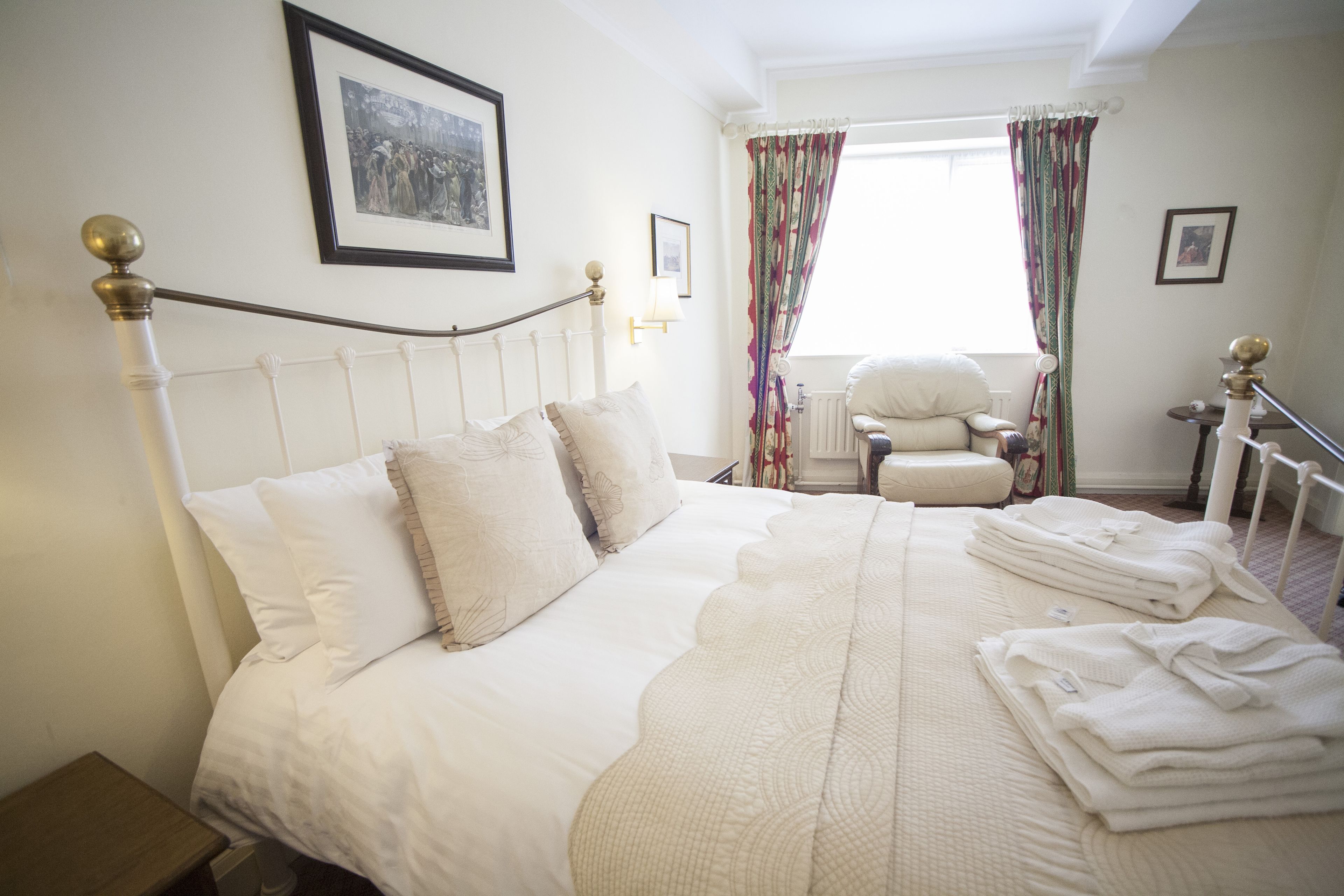 A neatly made bed with white linens and a decorative headboard, a chair, and a window with curtains.