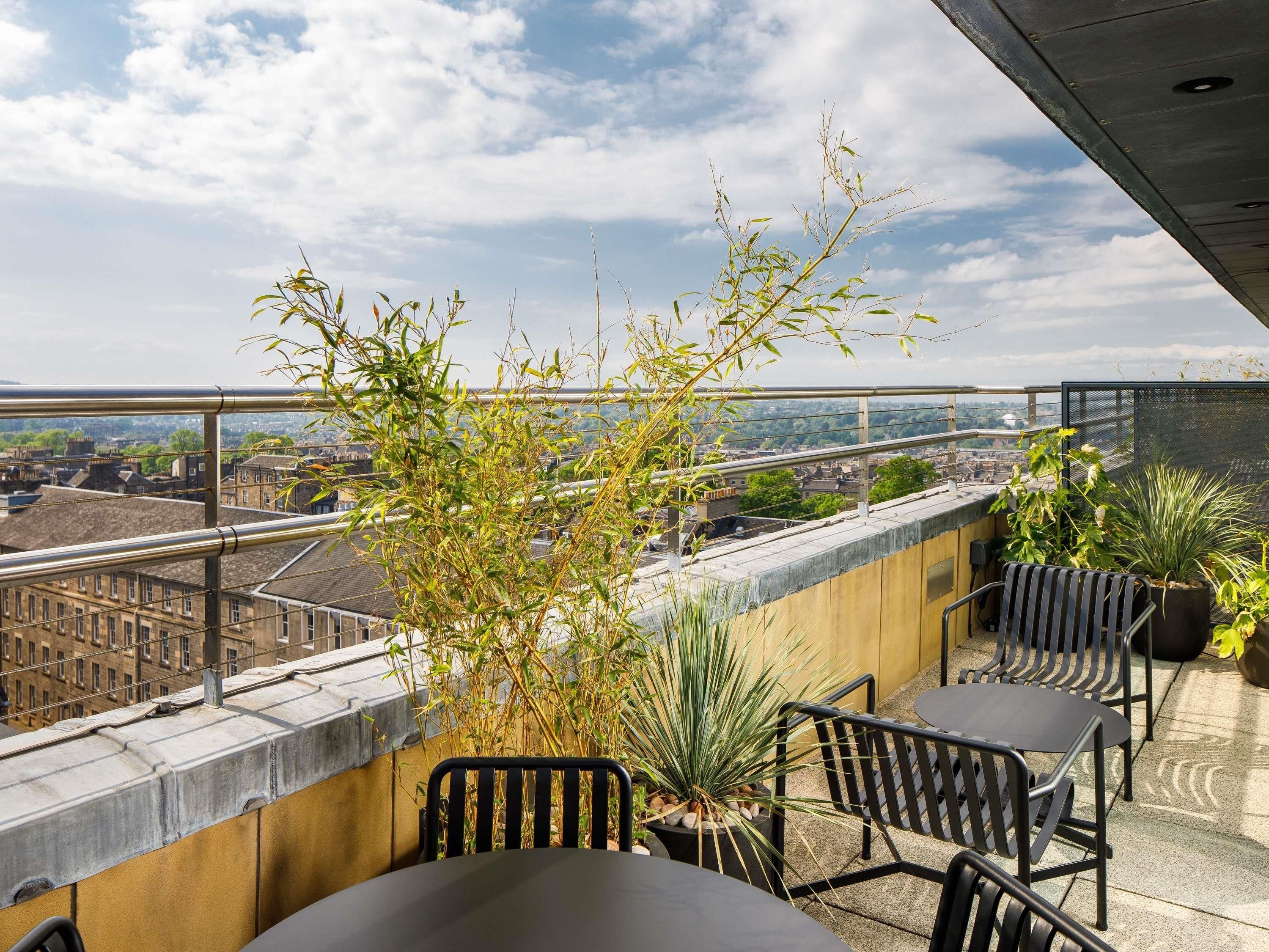 A rooftop terrace with a view of the city, featuring a table and chairs, potted plants, and a metal railing.