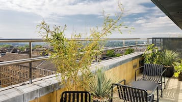 A rooftop terrace with a view of the city, featuring a table and chairs, potted plants, and a metal railing.