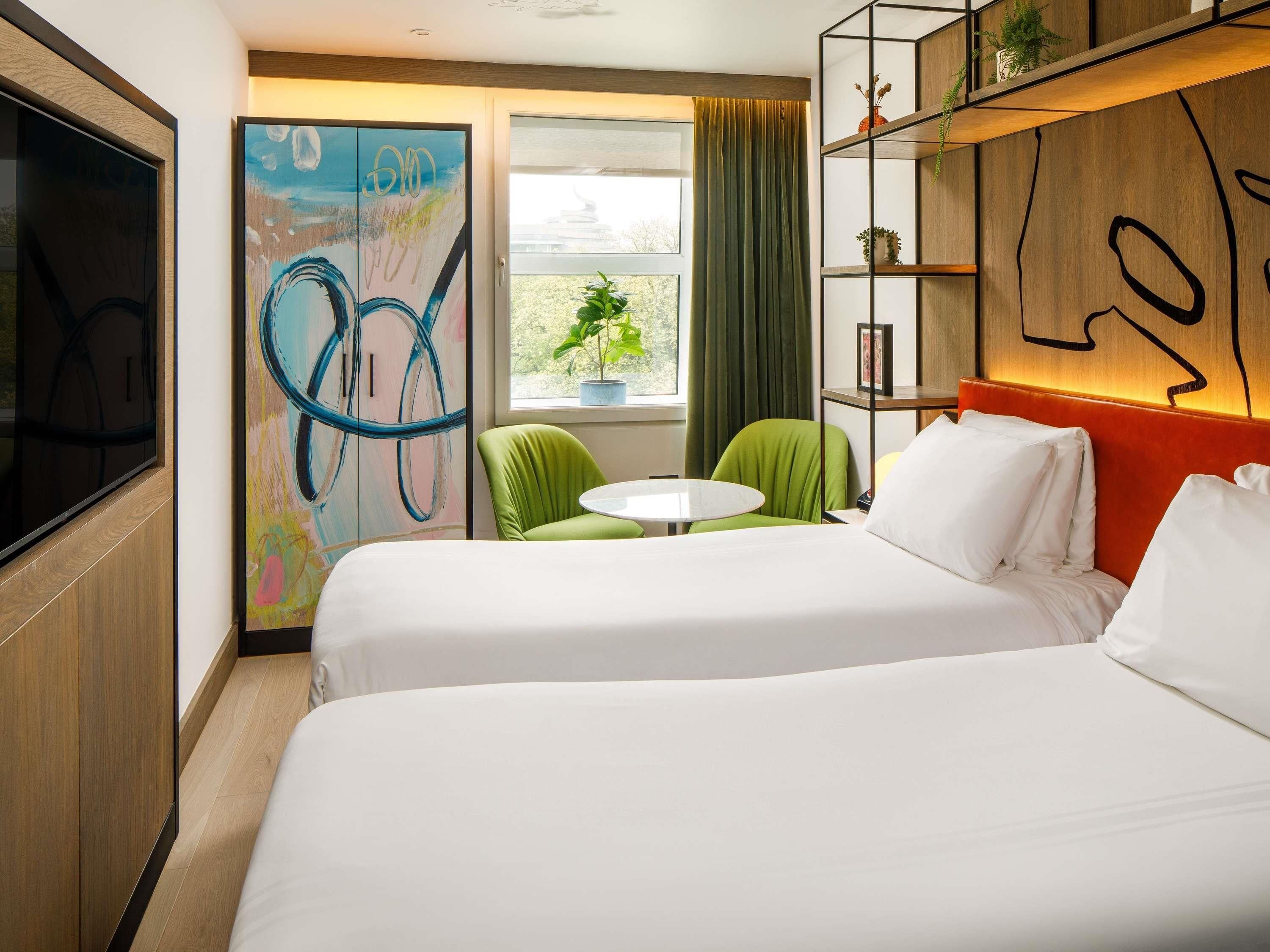 A hotel room with two beds, a TV, a painting, a green chair, a small table, and a window with curtains.