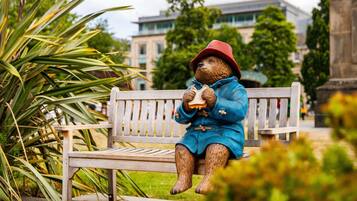 Una estatua de un oso con abrigo azul y sombrero rojo, sentado en un banco de parque.