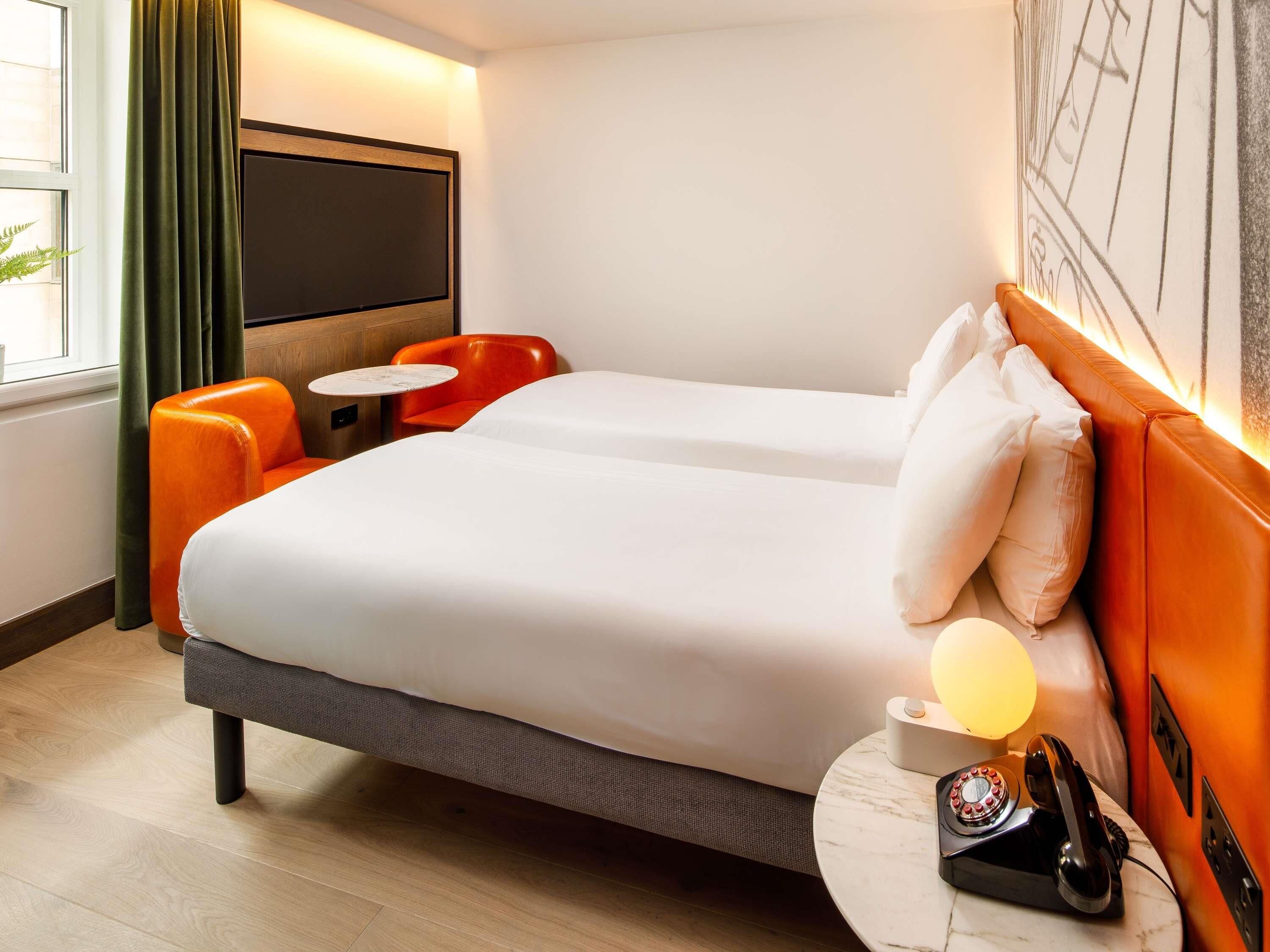 A hotel room with a bed, two chairs, a small table, a telephone, and a wall-mounted TV.