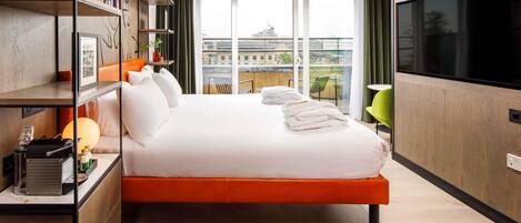A modern hotel room with a large bed, a TV, a bookshelf, and a view of the city.