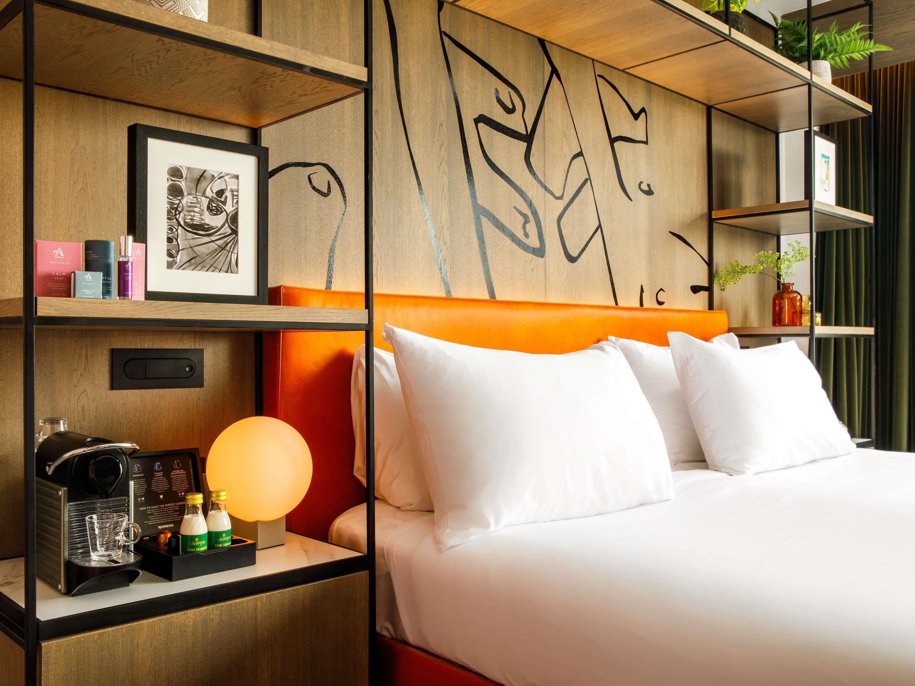 A hotel room with a bed, a nightstand with a coffee machine, a shelf with toiletries, and a wall with abstract drawings.