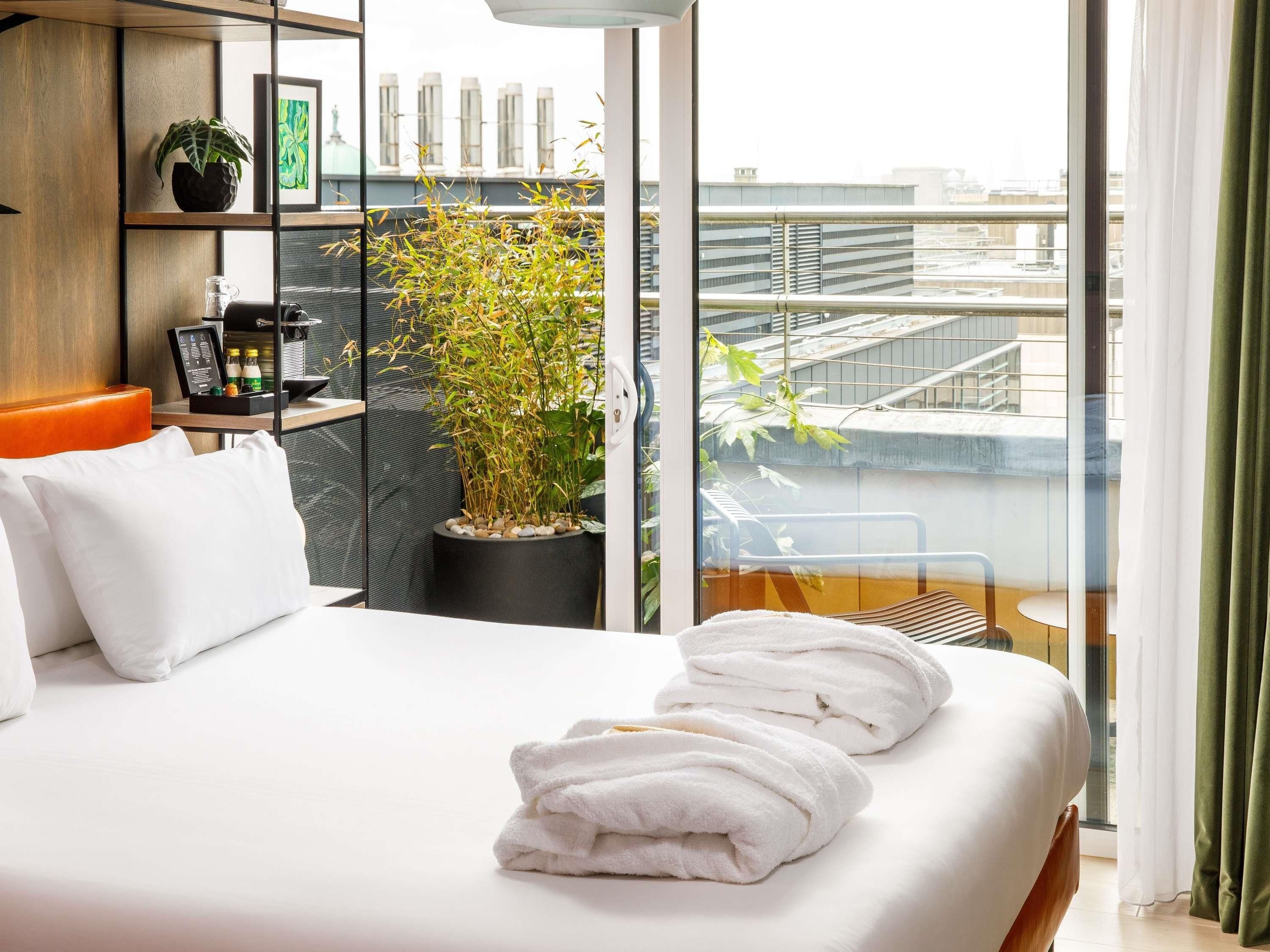 A hotel room with a bed, white pillows, a potted plant, a coffee machine, and a view of the city.