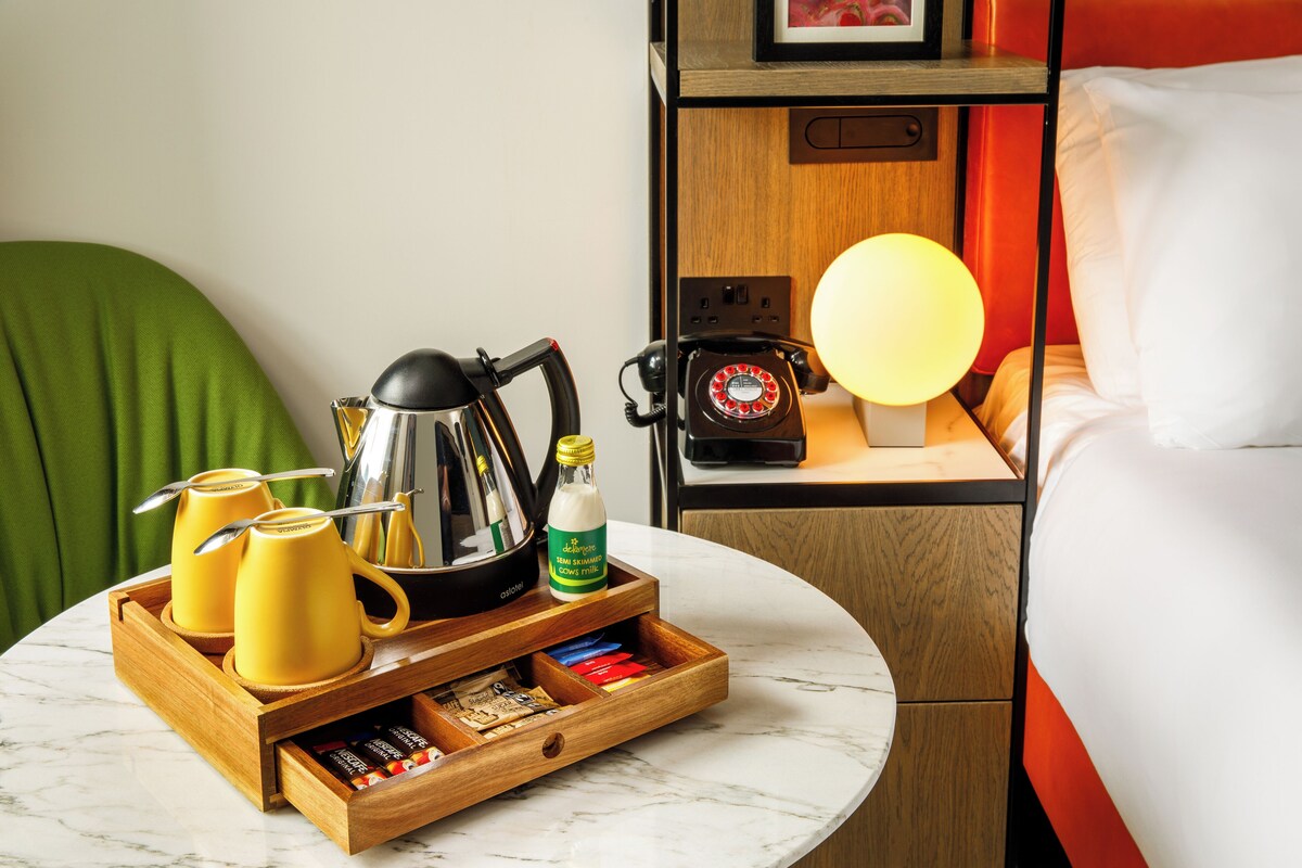 A neatly arranged hotel room with a wooden side table, a coffee maker, two yellow mugs, a bottle of milk, and a drawer with various items.
