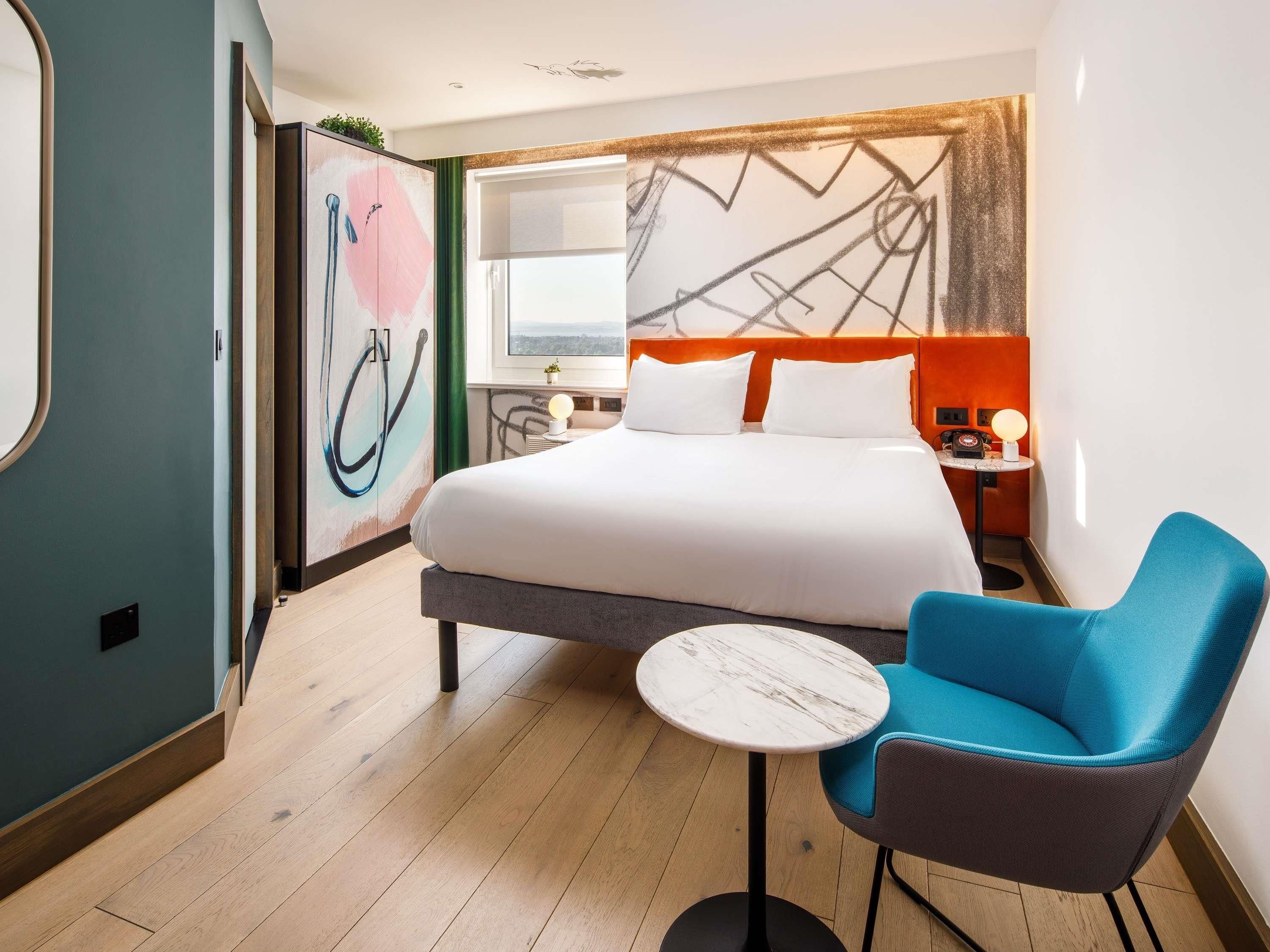 A hotel room with a bed, a small round table, a blue chair, and a wall with abstract art.