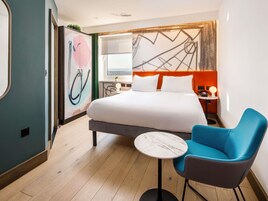A hotel room with a bed, a small round table, a blue chair, and a wall with abstract art.