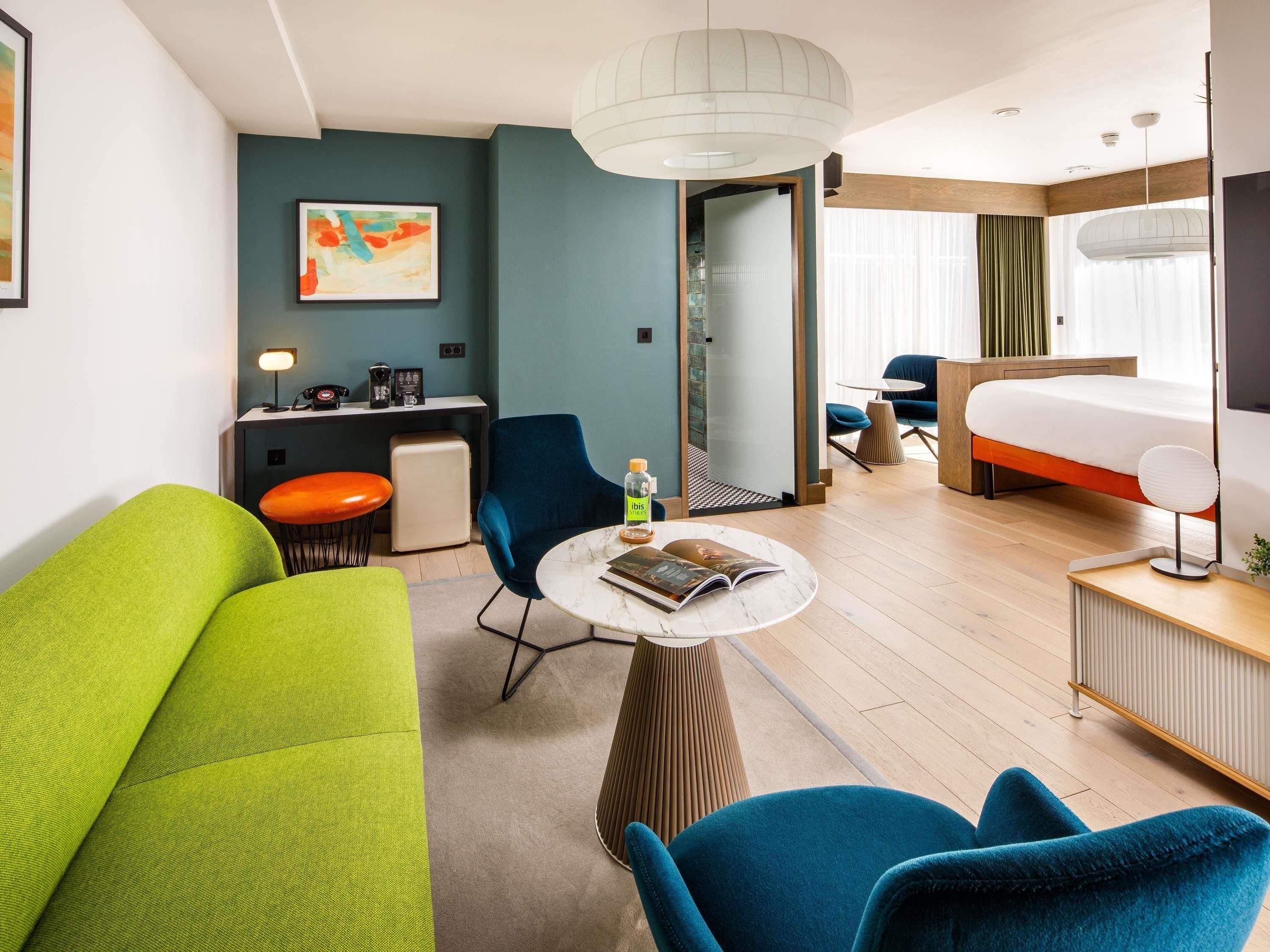A modern hotel room with a bed, seating area, and a desk with a lamp.