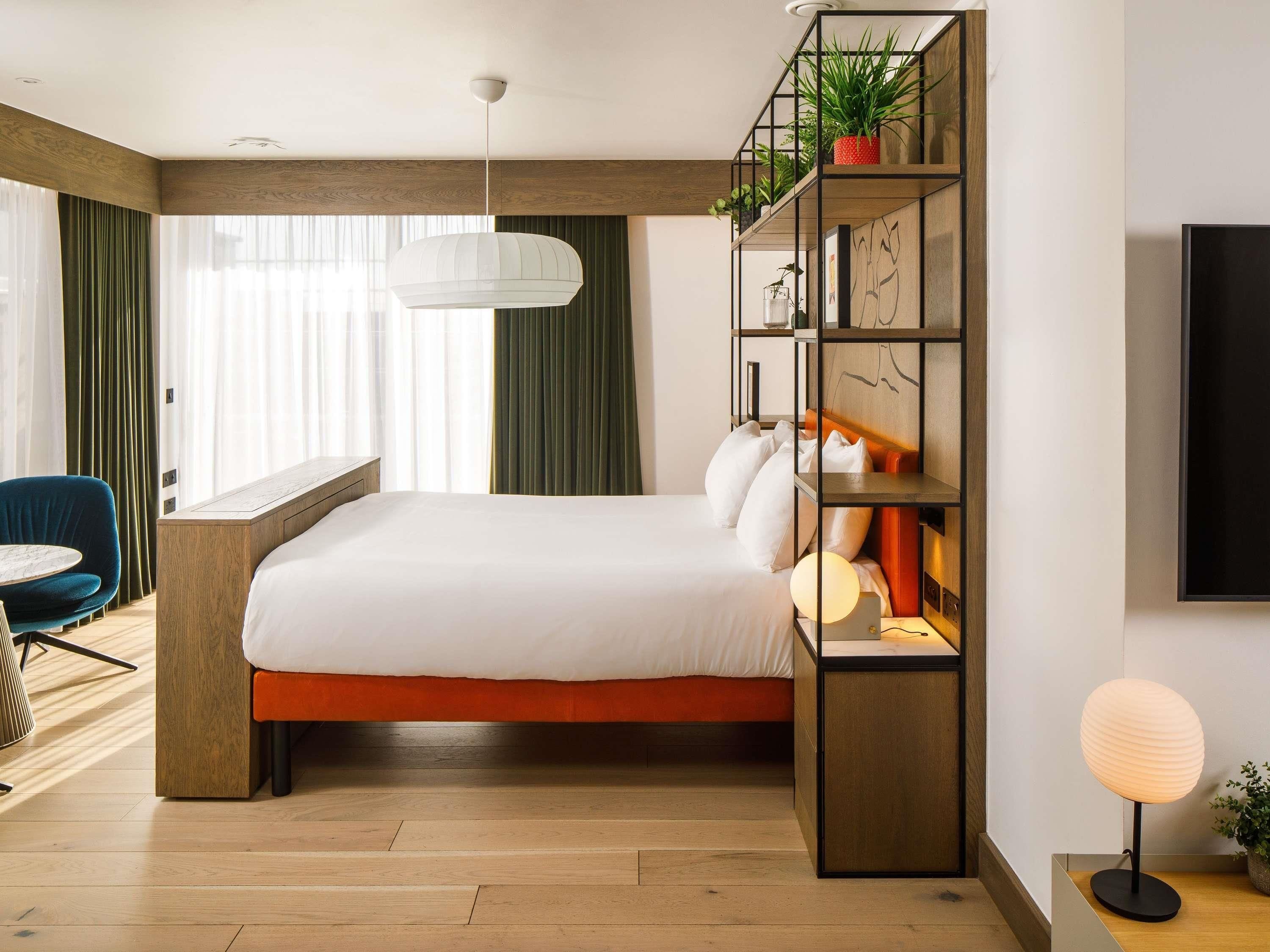 A modern hotel room with a large bed, a desk, a chair, a bookshelf, and a wall-mounted TV.