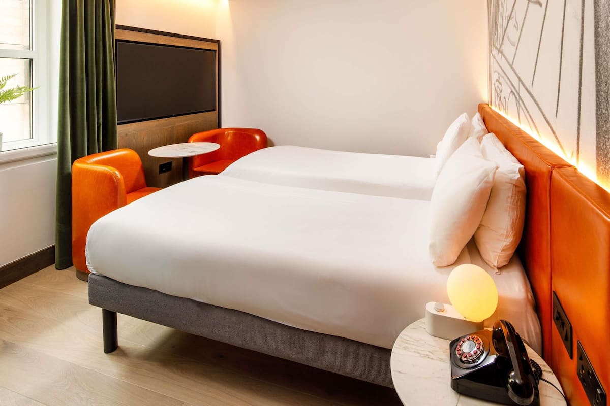 Une chambre d’hôtel comprenant un lit, deux chaises, une petite table, un téléphone et un téléviseur fixé au mur.