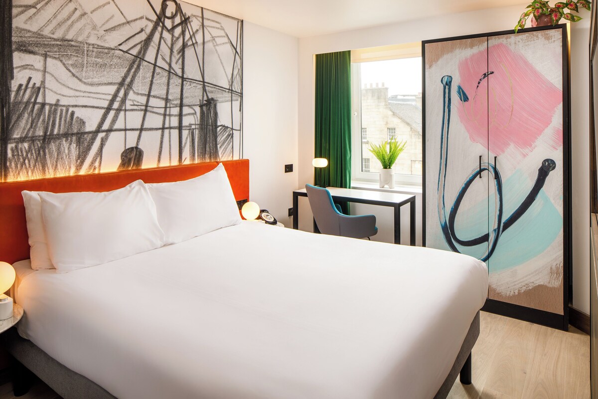 A hotel room with a large bed, a desk, a chair, a window with curtains, and a wall with abstract art.