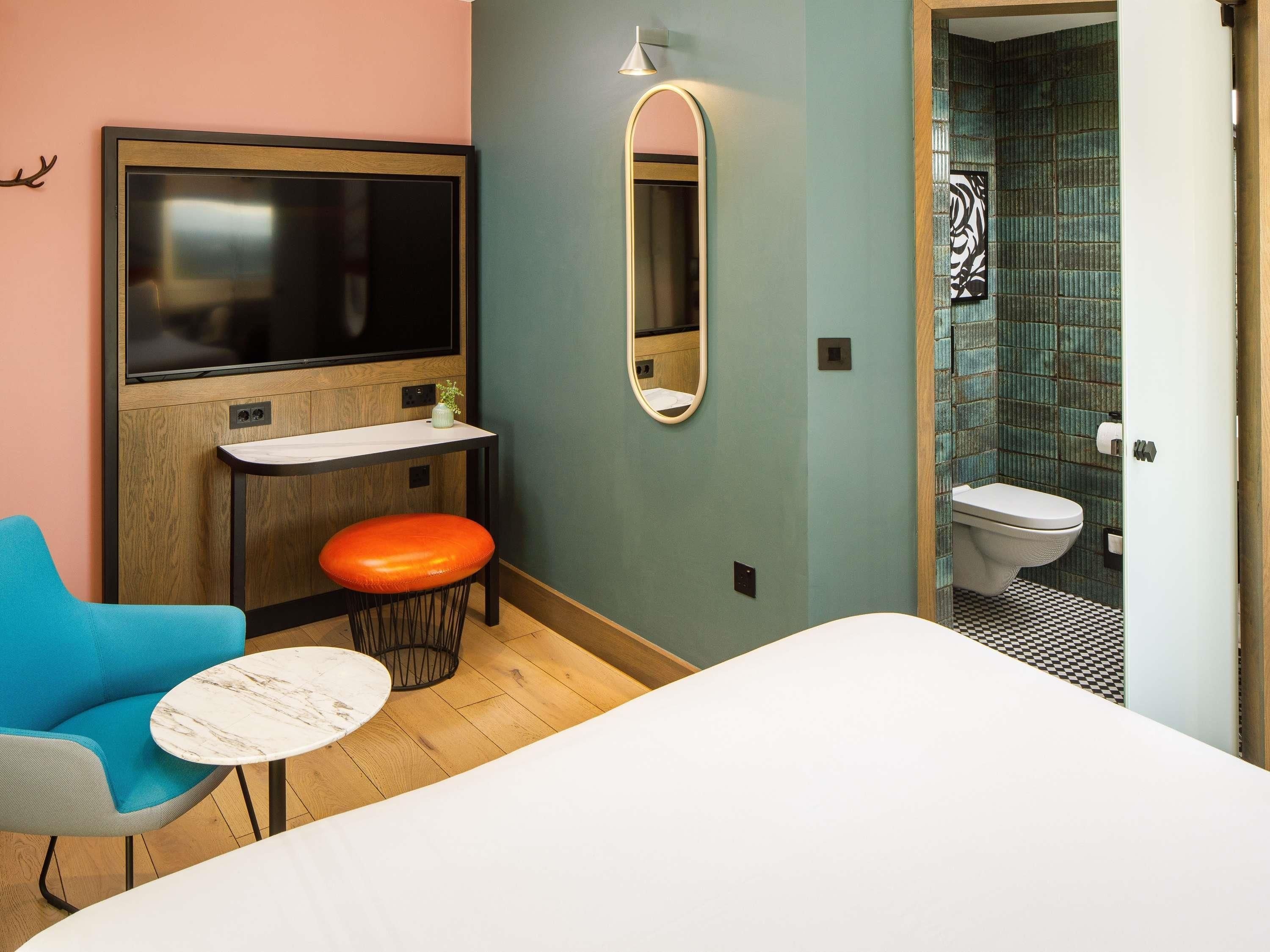 A hotel room with a bed, a seating area with a chair and table, a TV, a mirror, and a bathroom with a toilet and sink.