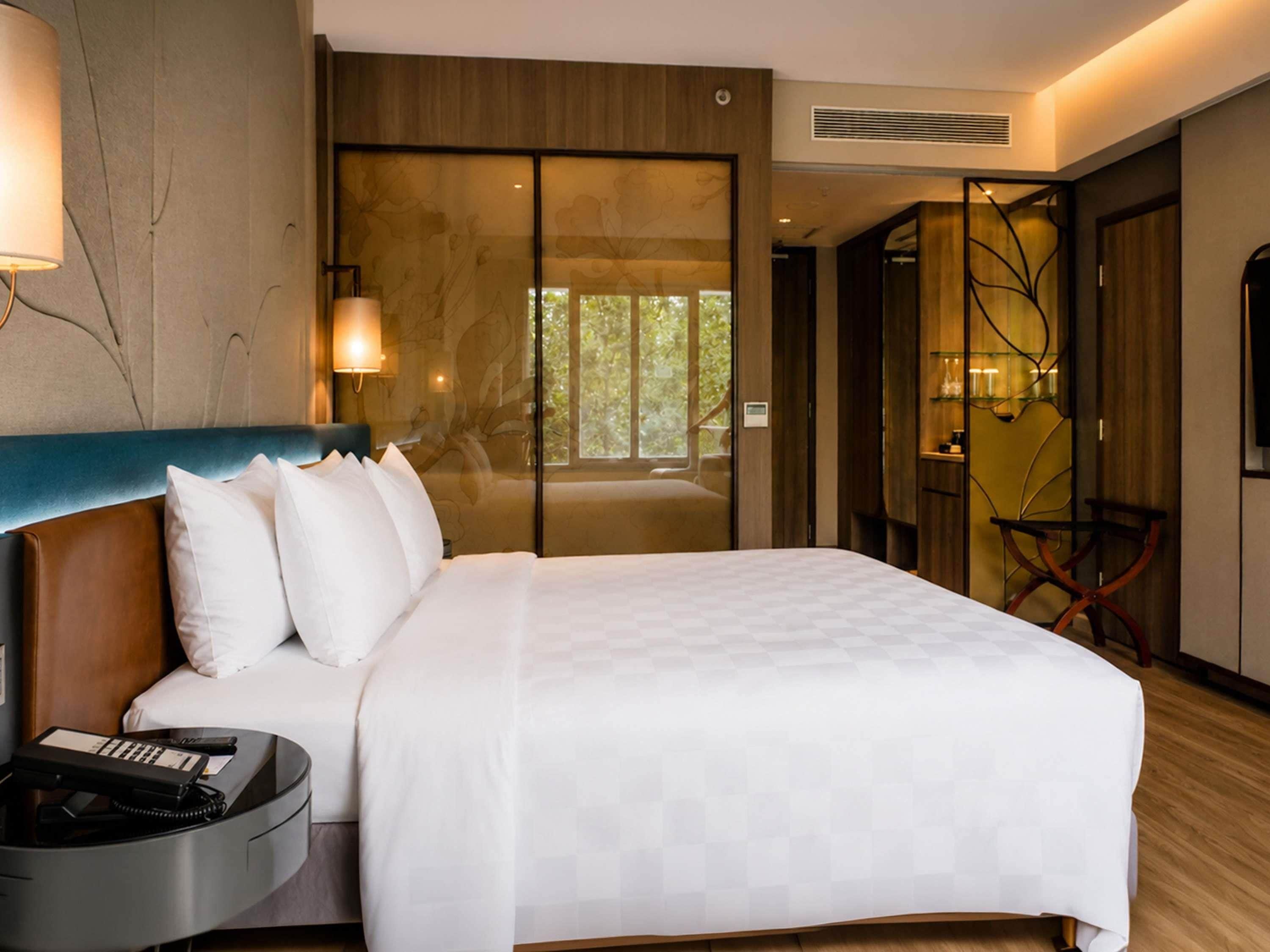 Une chambre d’hôtel avec un grand lit, une table de chevet avec un téléphone, une lampe fixée au mur et une porte coulissante donnant sur un balcon.