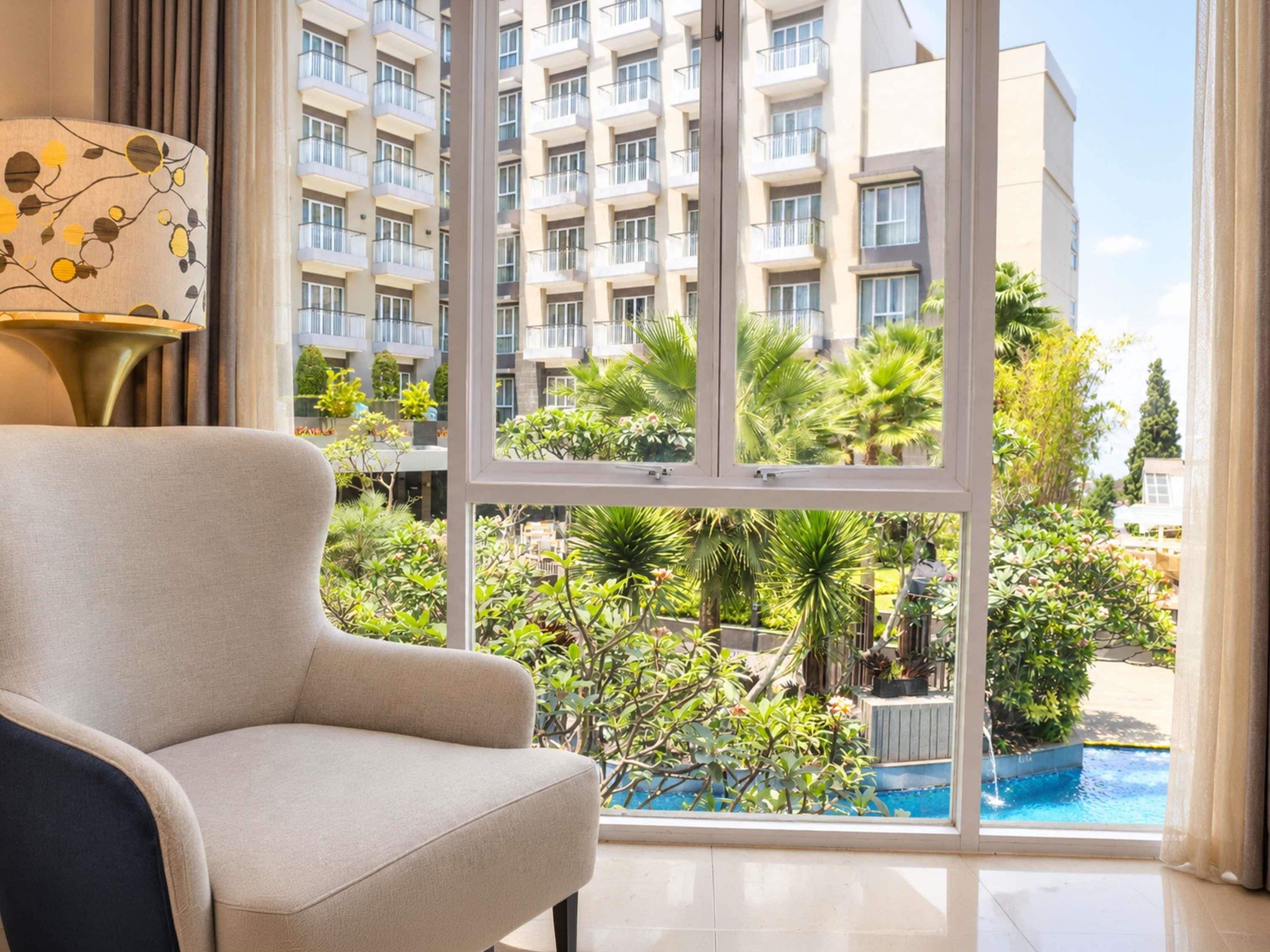 A modern hotel room with a large window, a sofa, and a view of a swimming pool and greenery.
