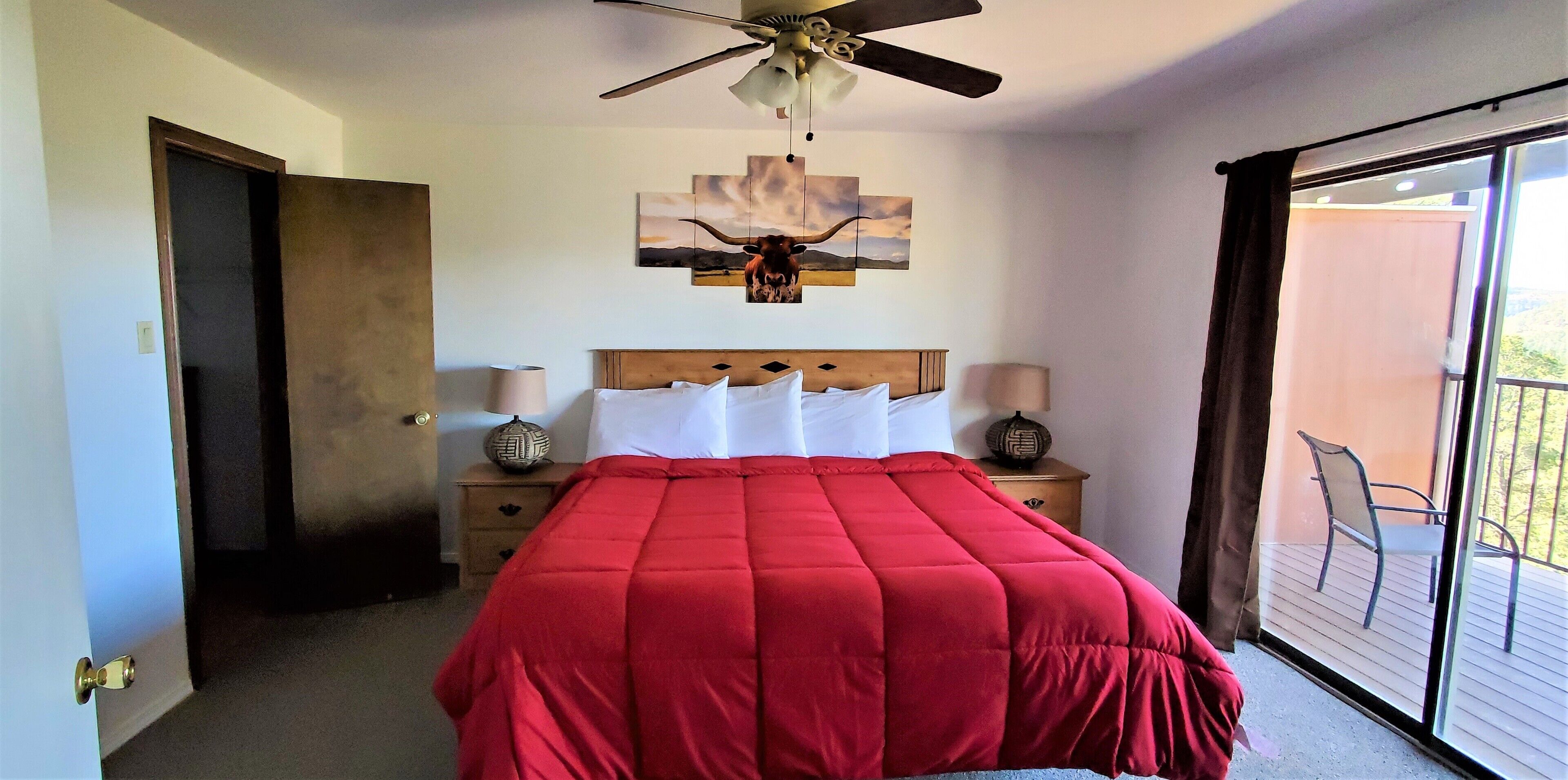 A bedroom with a large bed, a ceiling fan, a nightstand with a lamp, and a view of the outside balcony.