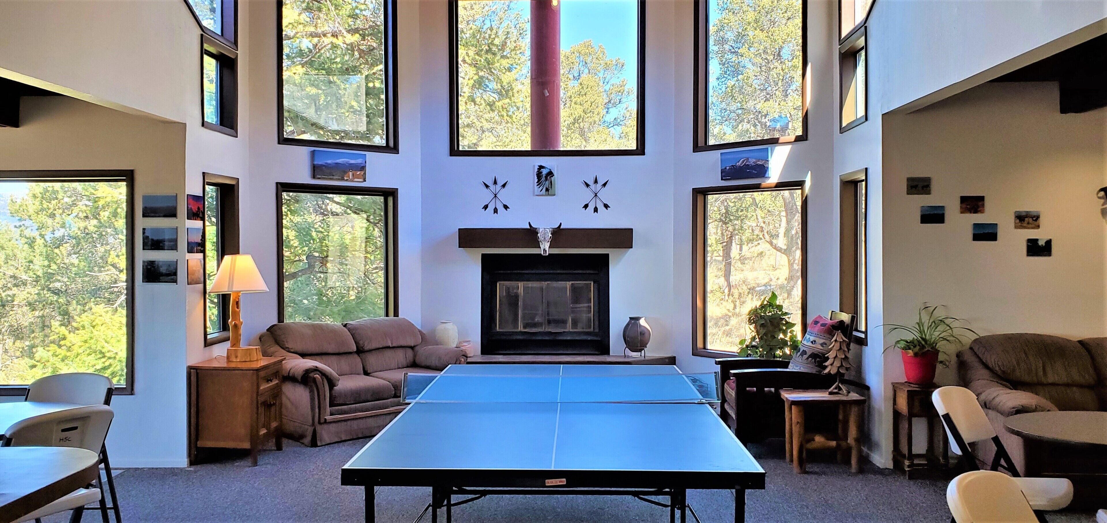Ein geräumiges Wohnzimmer mit Tischtennisplatte, Kamin und großen Fenstern mit Blick auf Bäume.