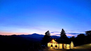 A small, lit cabin with a wooden exterior, surrounded by trees and mountains in the background. - High Sierra Condominium (Ruidoso)