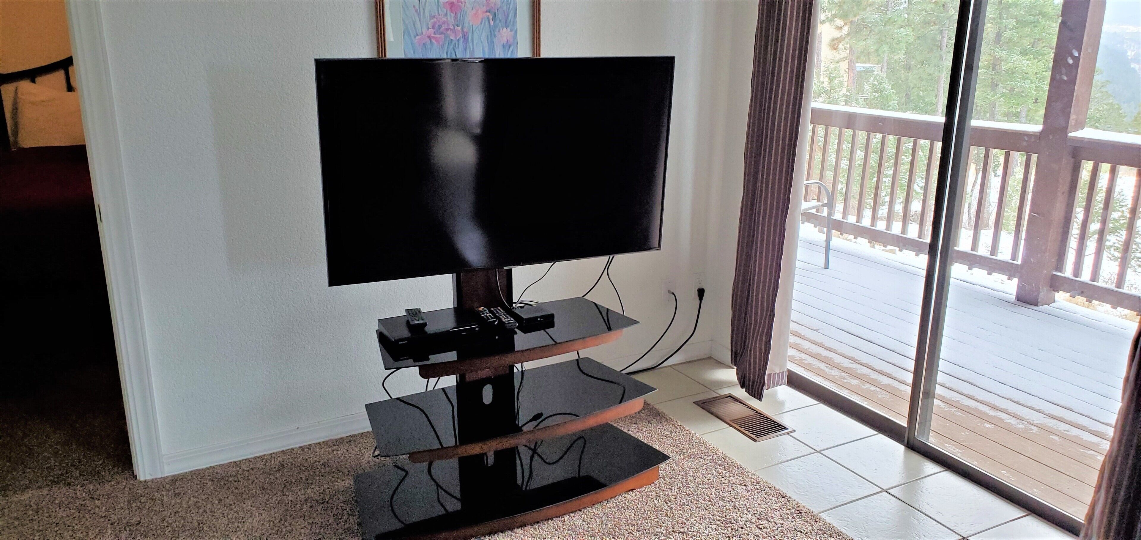 Televisão de tela plana sobre suporte de madeira em um quarto com uma janela ampla e vista para uma varanda.