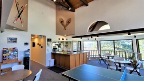 A spacious indoor area with a ping pong table, seating, a Coca-Cola vending machine, and a view through large windows. - High Sierra Condominium (Ruidoso)