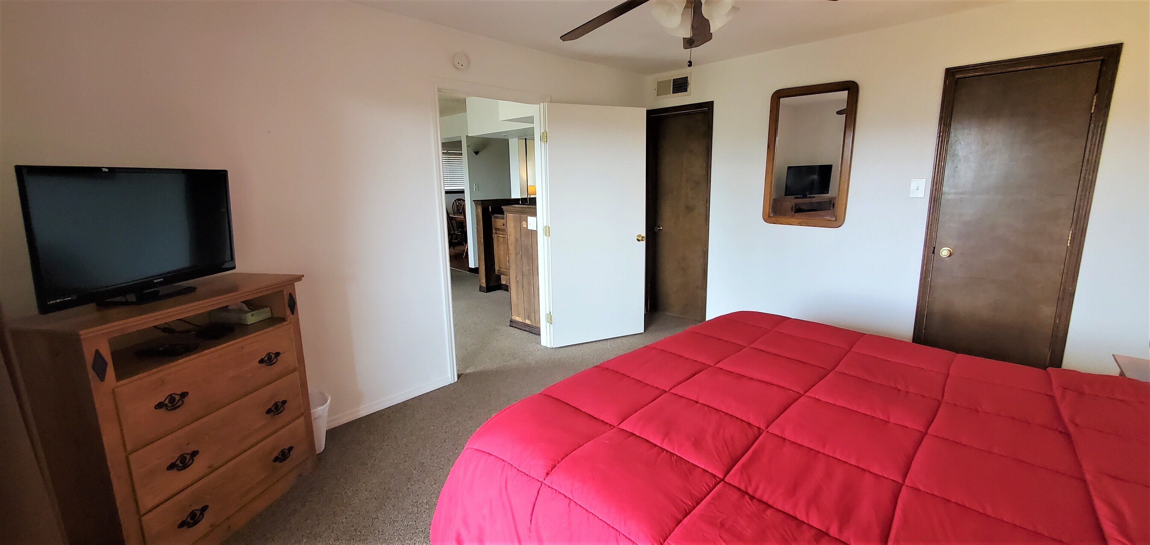 Une chambre avec une couvre-lit rouge, un téléviseur posé sur une commode en bois et un miroir accroché au mur.
