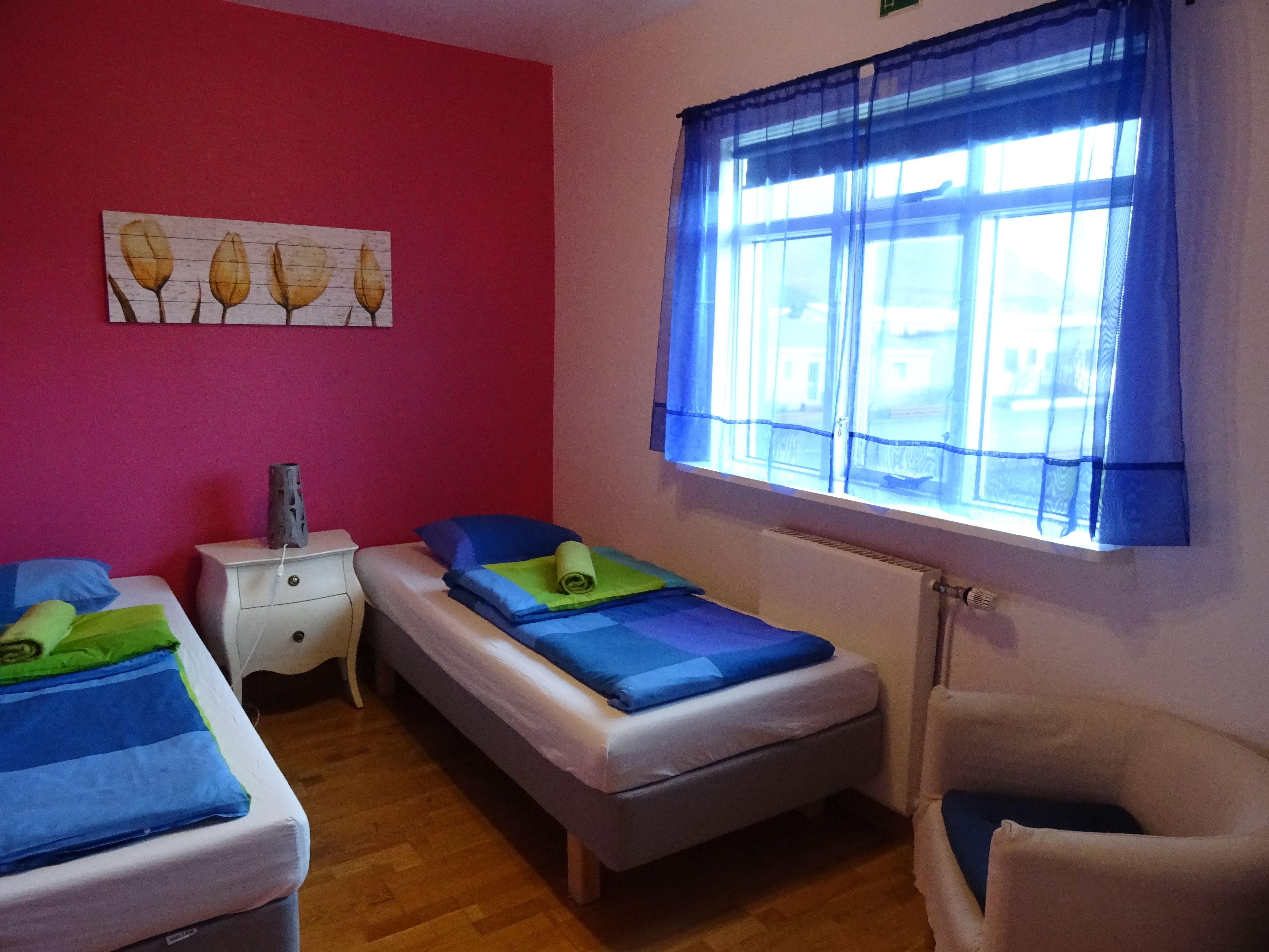A bedroom with two beds, a nightstand, a chair, a window with blue curtains, and a painting on the wall.
