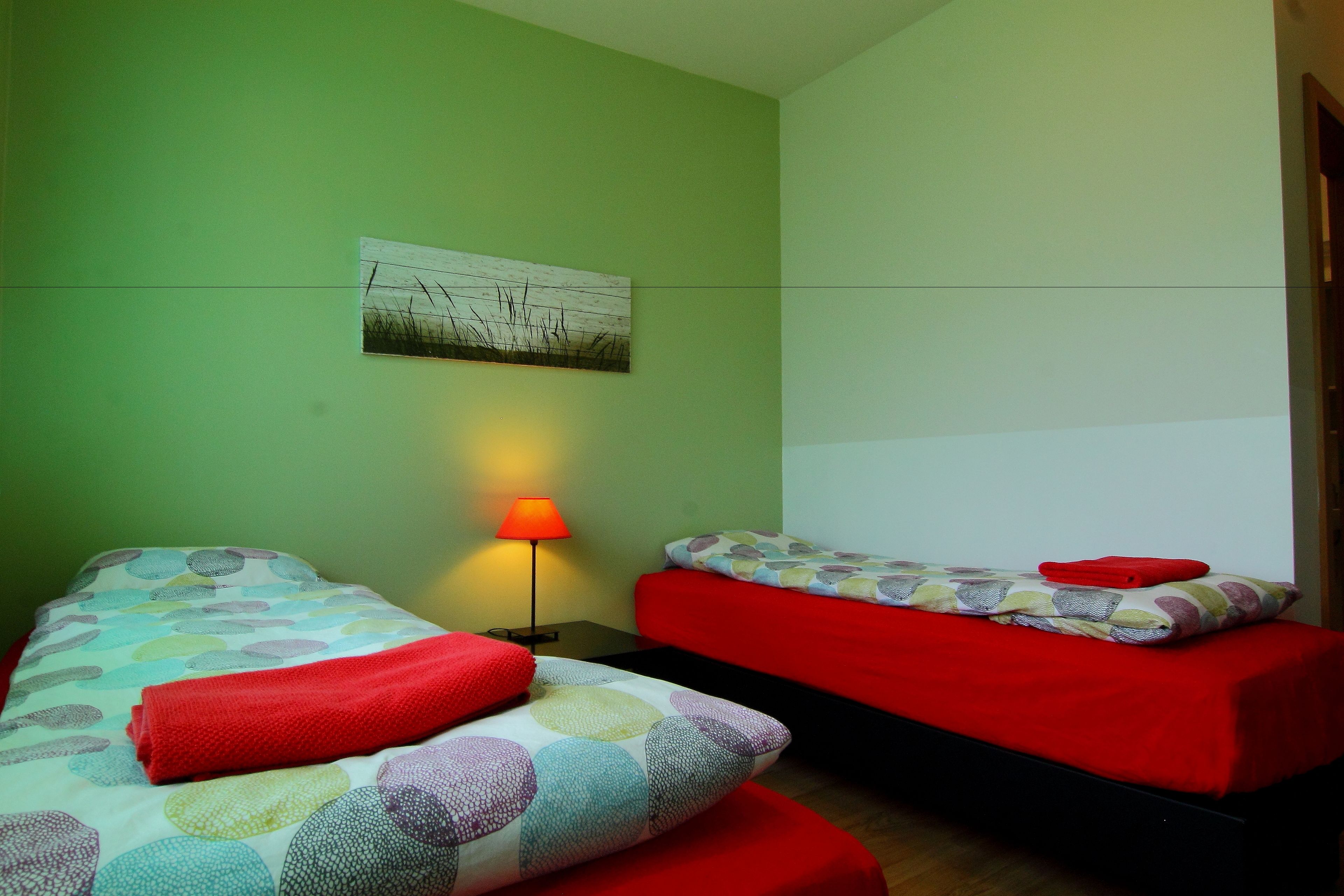 A room with two beds, each with a red blanket, a lamp, and a wall-mounted picture.