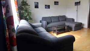 A living room with a grey sectional sofa, a small wooden coffee table, and a floor lamp. There are three framed pictures on the wall and a potted plant in the corner. - Grundarfjördur Hostel (Grundarfjörður)