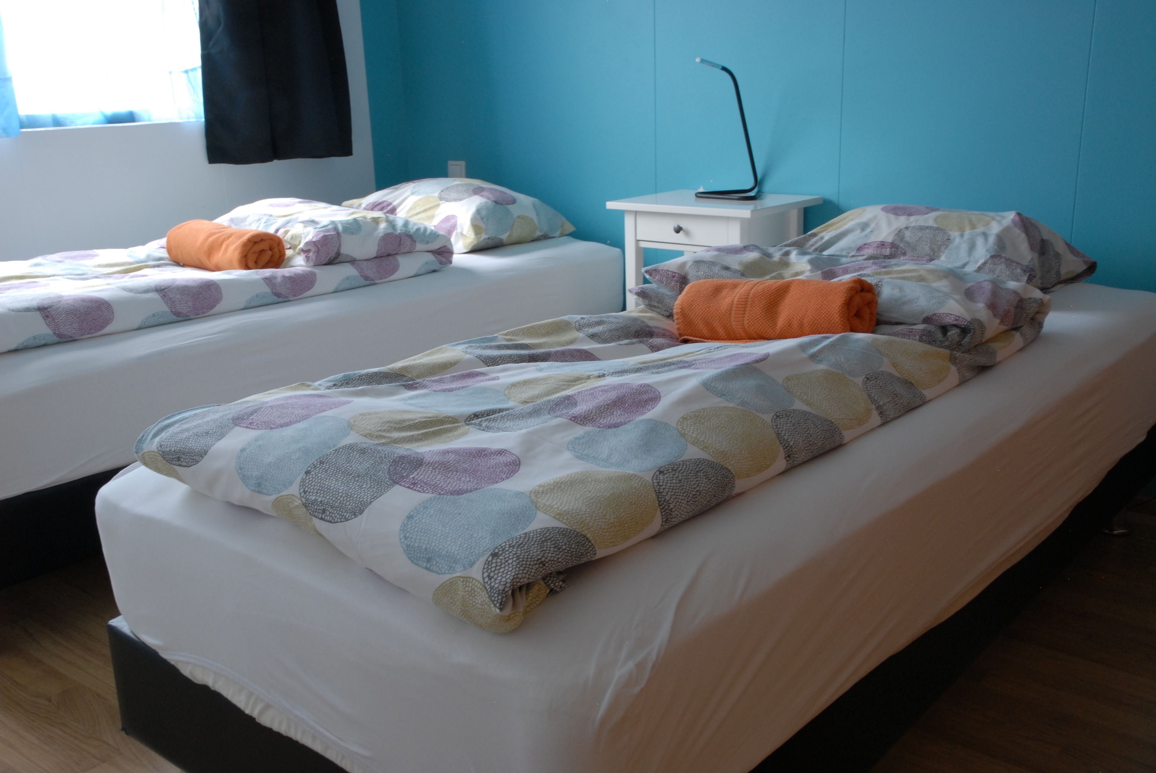 Hotellihuone, jossa on kaksi erillistä sänkyä, jokaisessa kuviollinen peitto ja tyynyt, yöpöytä lampun kanssa sekä ikkuna, jossa on verhot.