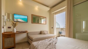 Habitación de hotel con cama, sofá, escritorio con un florero de flores secas, televisor de pantalla plana y un ventanal con vista a la playa.