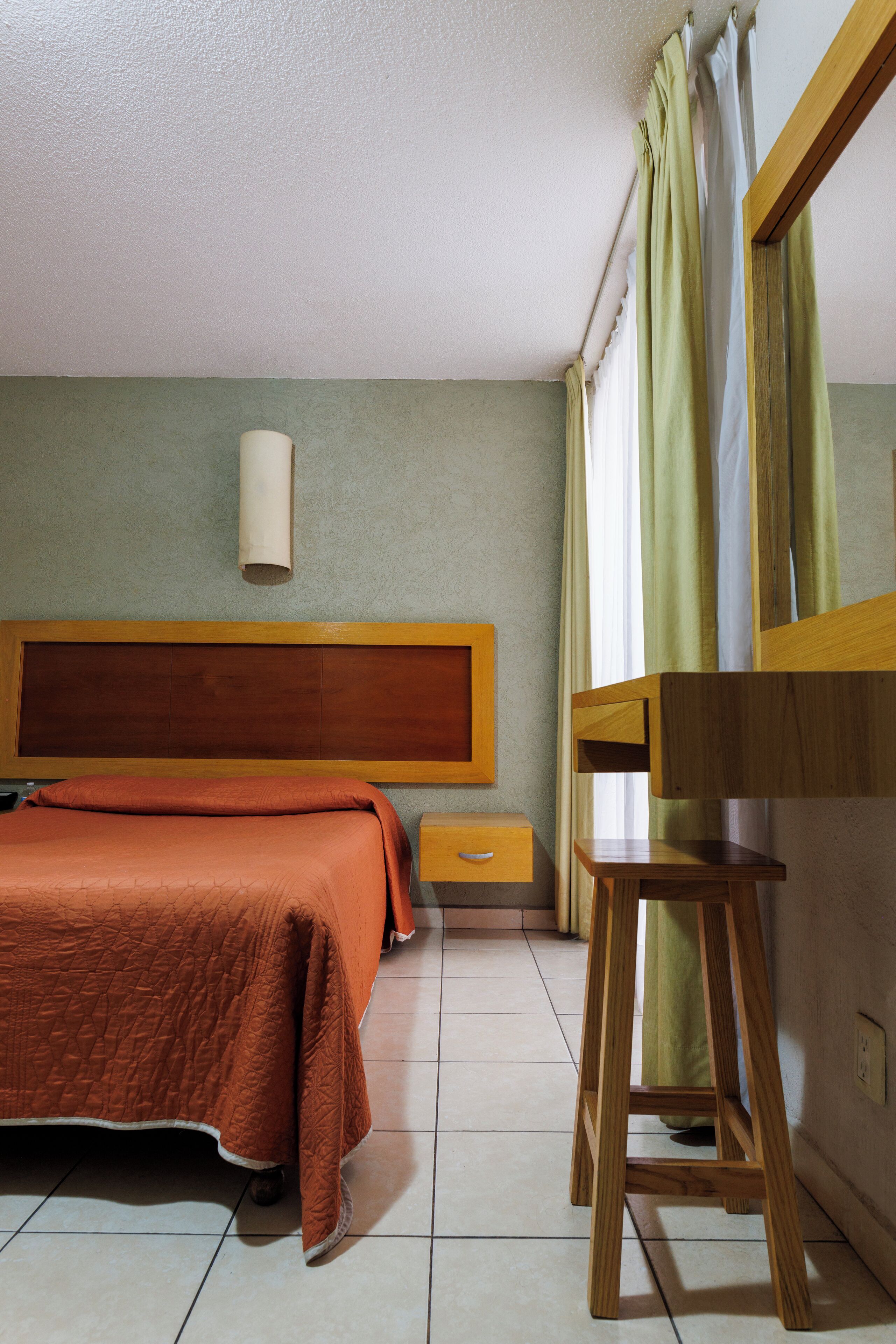 Een hotelkamer met een bed, een houten nachtkastje, een spiegel en een raam met gordijnen.