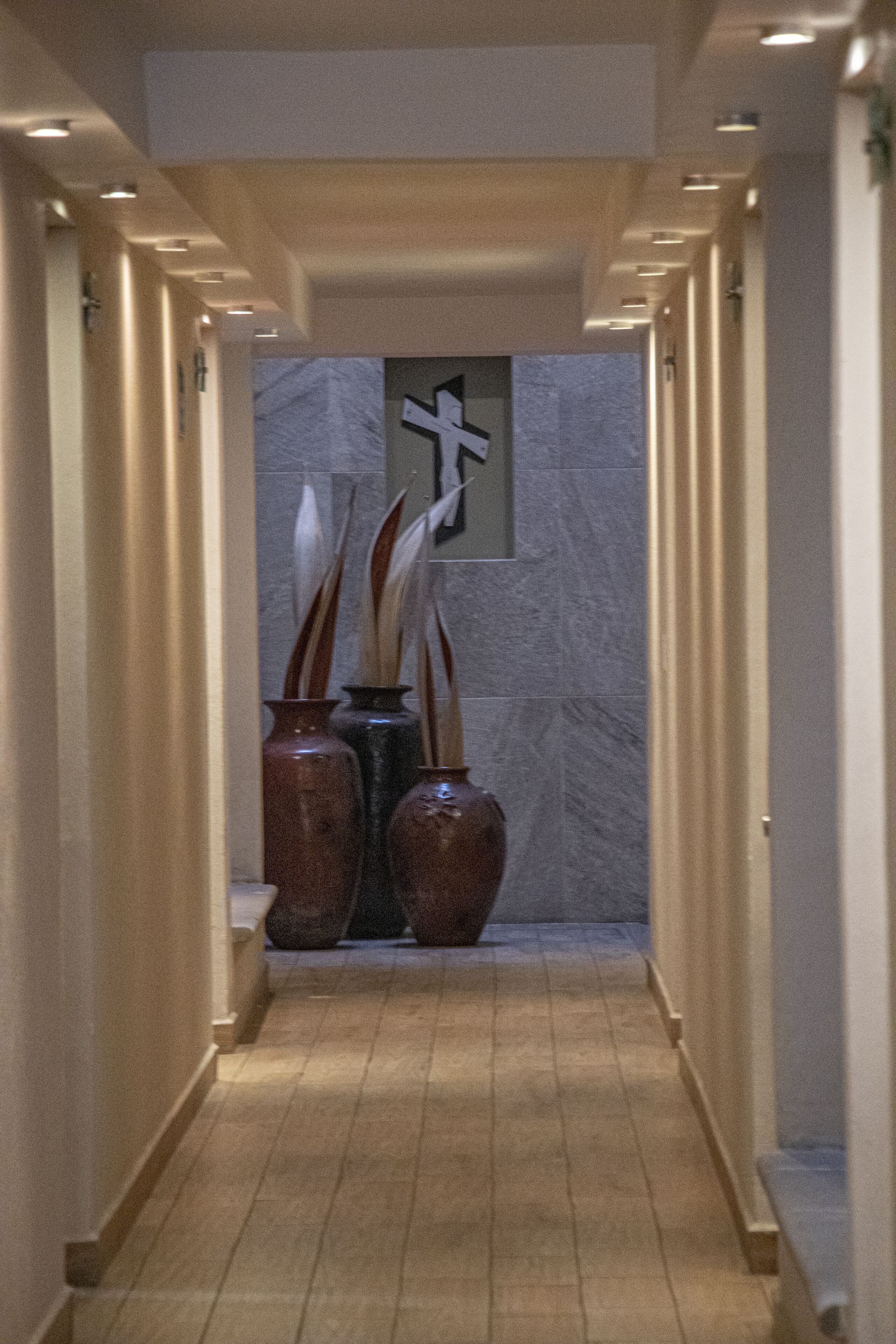 A hallway with a marble wall and a cross decoration. There are two large vases with tall, slender plants. The floor is wooden, and the walls are beige.