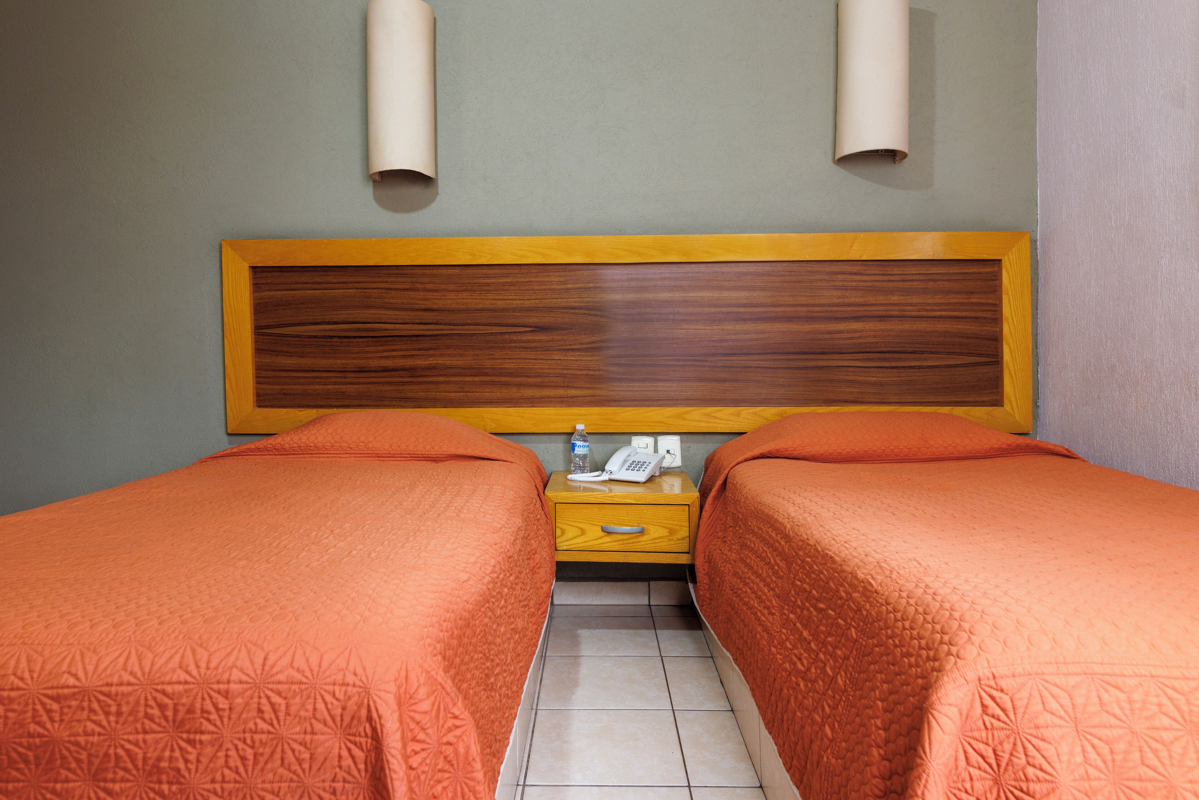 A hotel room with two beds, a wooden headboard, a nightstand with a phone and a bottle, and a light switch on the wall.