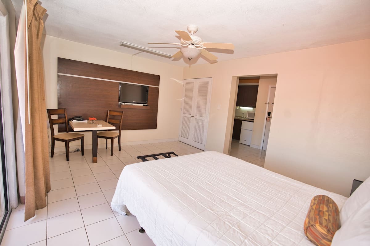 A hotel room with a bed, a desk with chairs, a ceiling fan, a TV mounted on the wall, and a door leading to a bathroom.