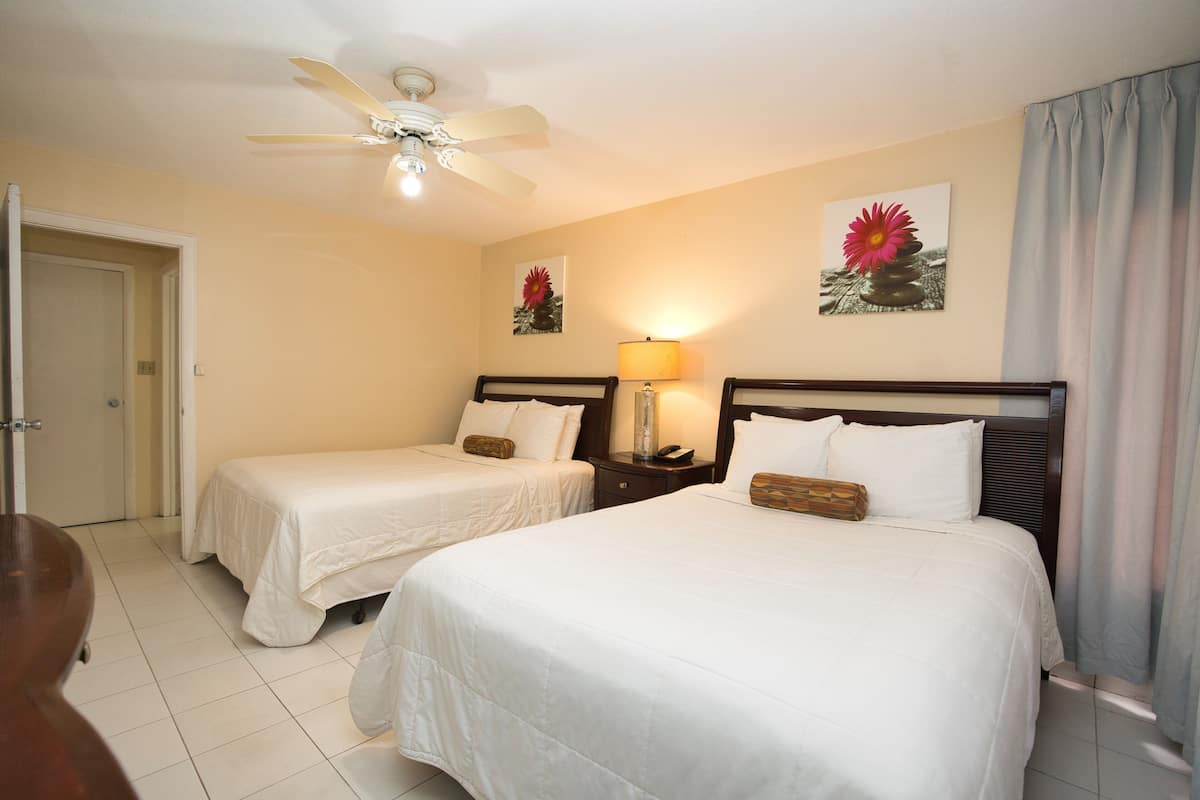 A hotel room with two single beds, a ceiling fan, a nightstand with a lamp, and artwork on the walls.