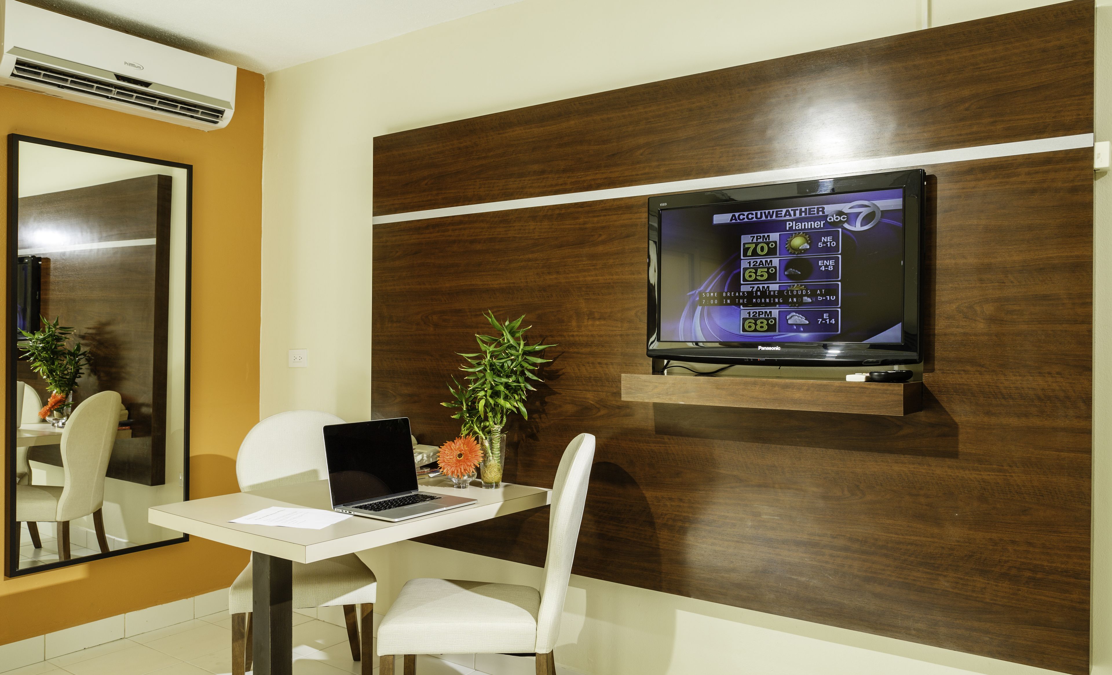 Habitación de hotel con pared decorativa de madera, televisor montado mostrando el pronóstico del tiempo, una computadora portátil, una mesita y una silla.