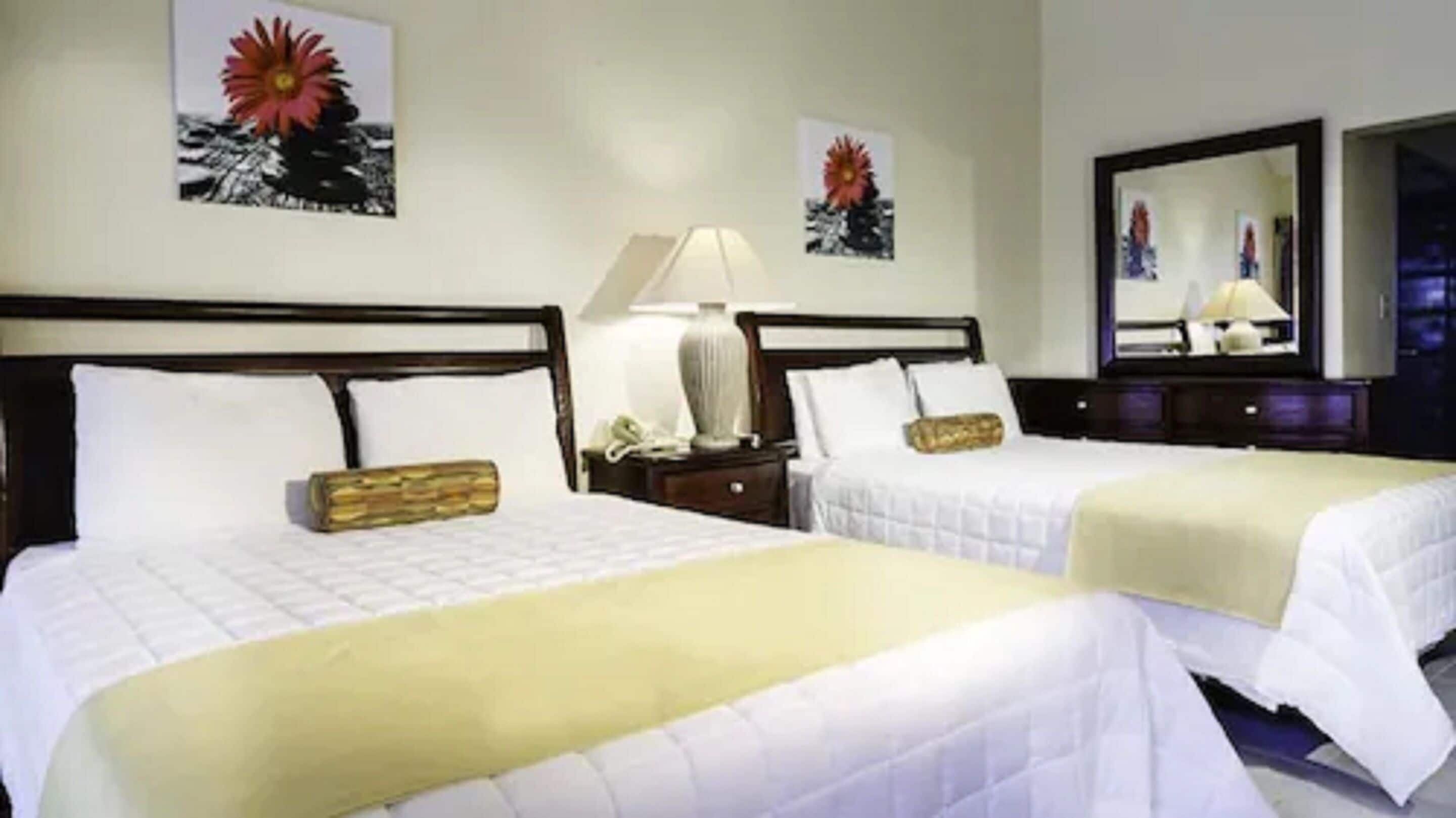 A hotel room with two beds, each with white bedding and a wooden headboard. There are two bedside tables with lamps and a vase on one. The walls have framed pictures of flowers.
