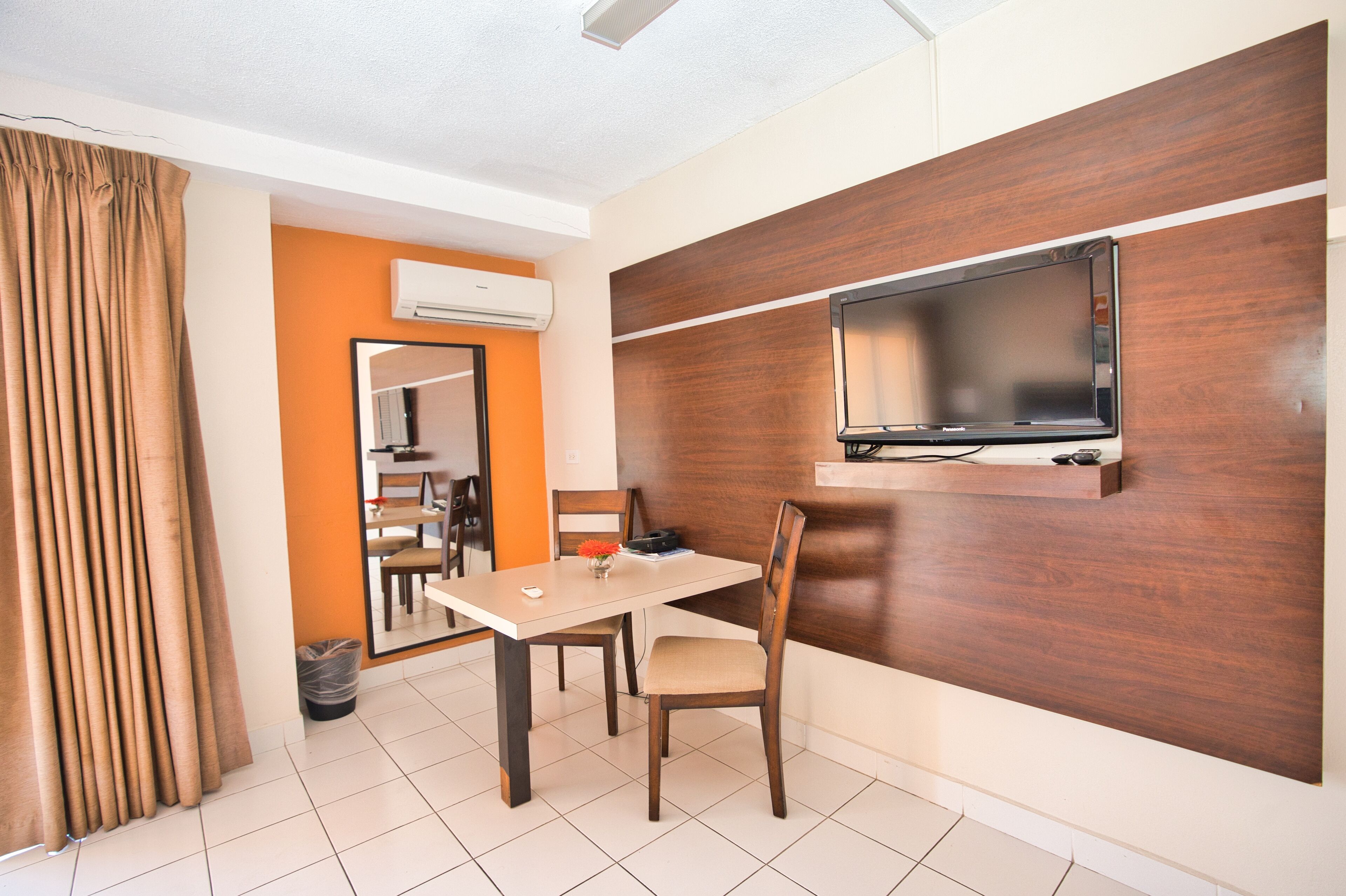 A hotel room with a dining area, a TV mounted on a wooden wall, and an air conditioning unit on the wall.