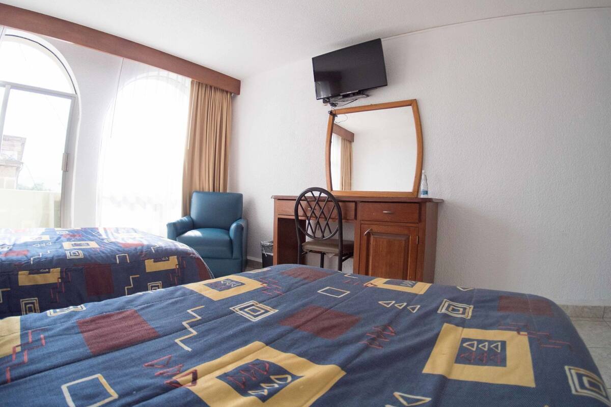 Hotellihuone, jossa on kaksi sänkyä, työpöytä tuolilla, seinälle asennettu televisio ja ikkuna, jossa on verhot.