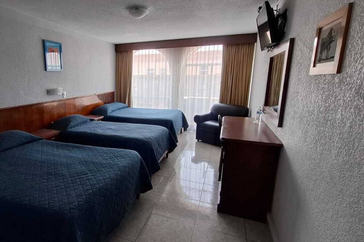 Hotellihuone, jossa on kolme sänkyä, työpöytä, tuoli ja ikkuna, jossa on verhot.