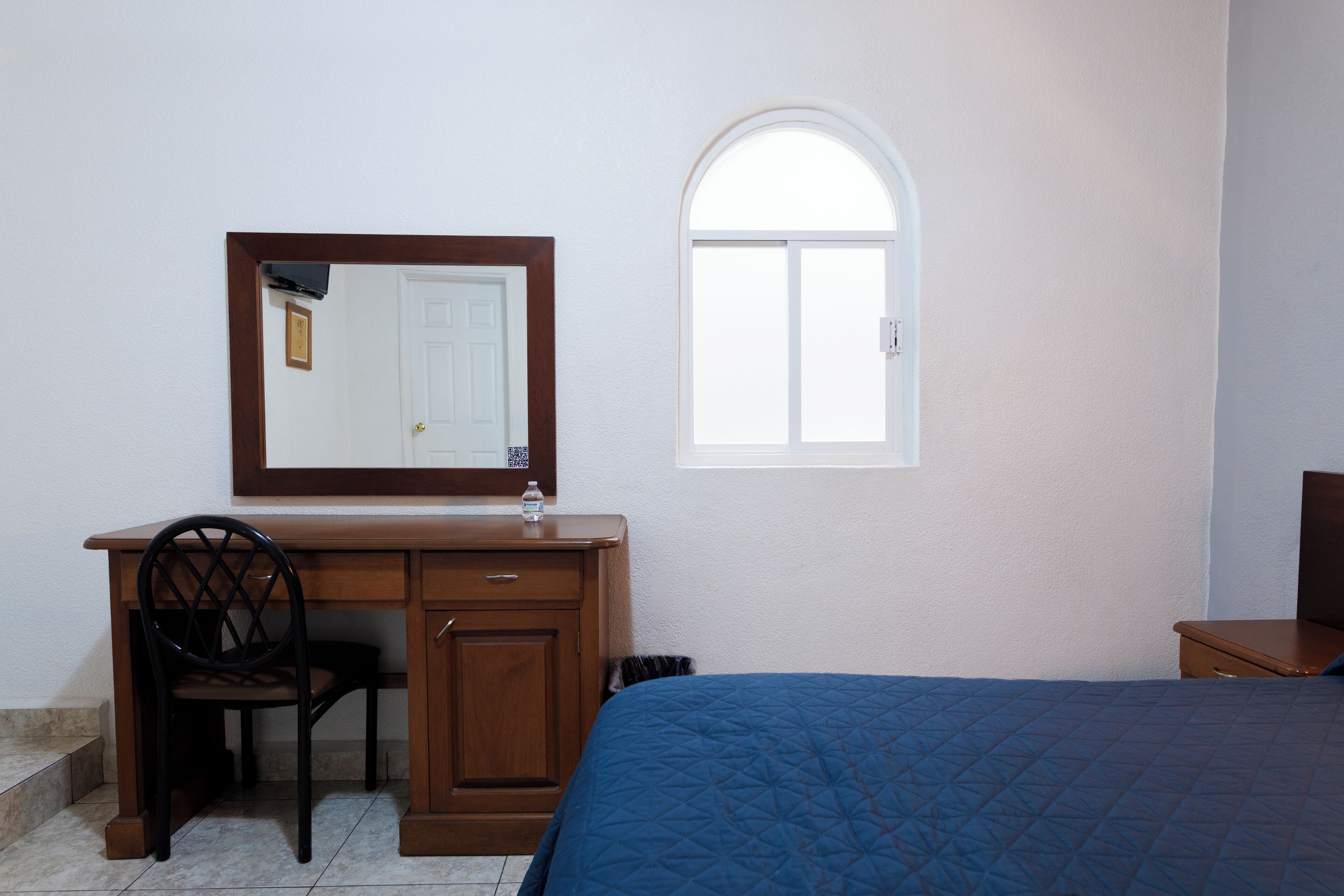 Un dormitorio con cama, escritorio, silla, ventana pequeña y puerta.