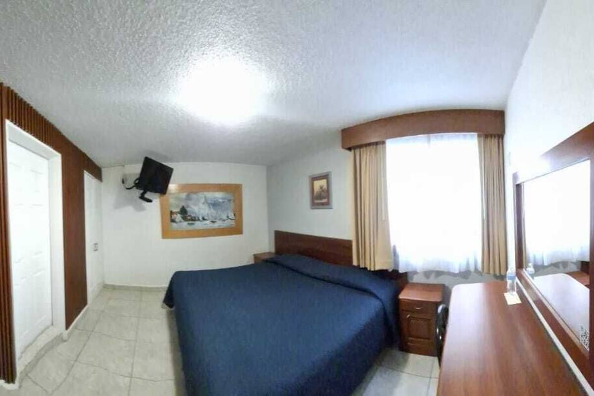 Hotellihuone, jossa on sänky, seinälle asennettu televisio, puuovi ja ikkuna, jossa on verhot.