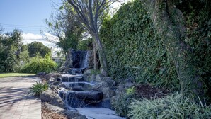 Un jardín ajardinado con una fuente que cae en cascada, un sendero pavimentado y abundante vegetación.