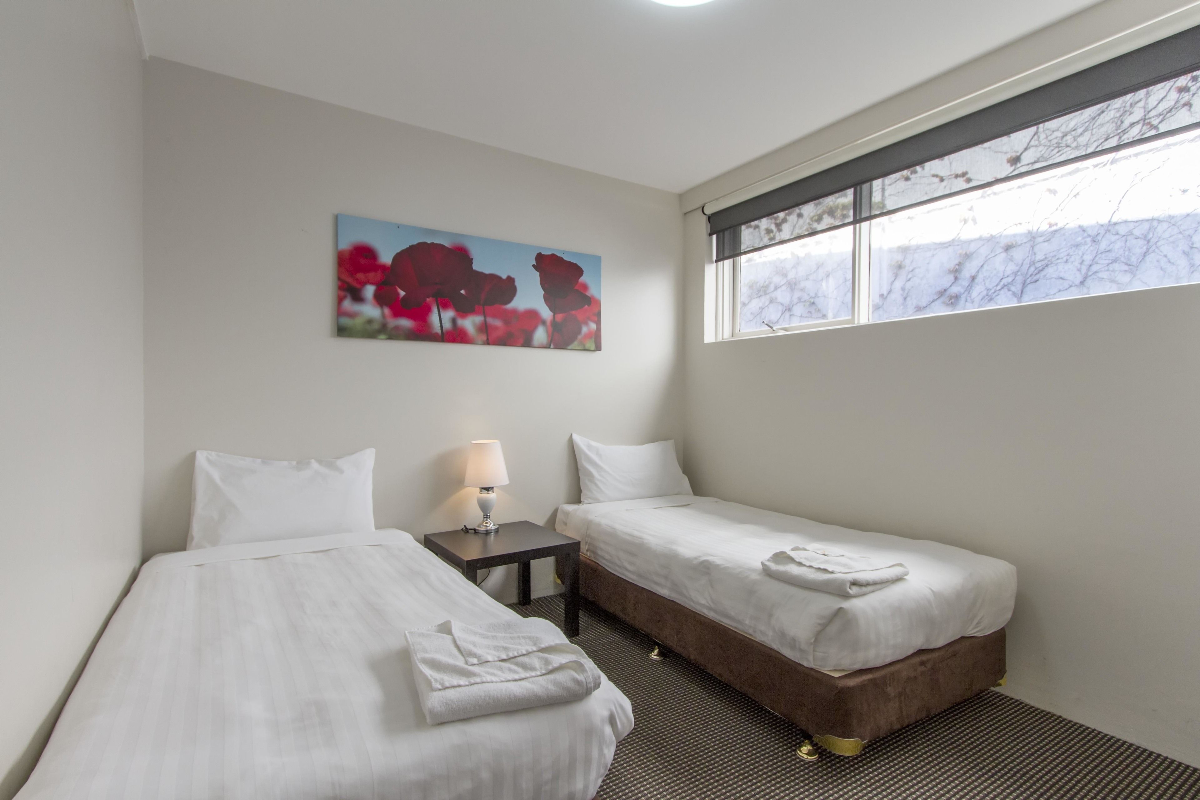Habitación de hotel con dos camas individuales, una mesita con lámpara, un cuadro de flores rojas y ventana con vista a árboles.