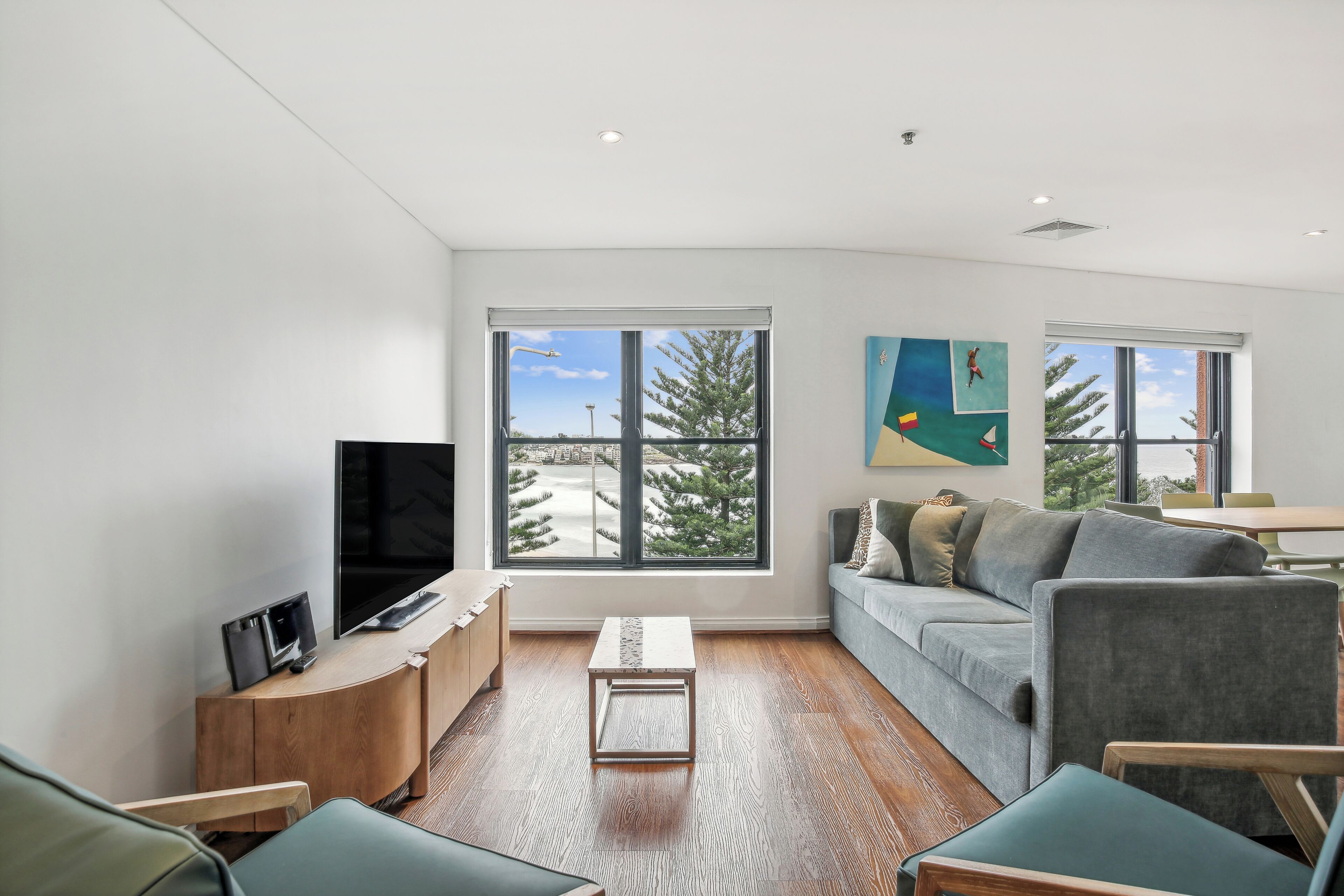 A modern living room with a large TV, wooden furniture, a grey sofa, and large windows offering a view of trees and a building.