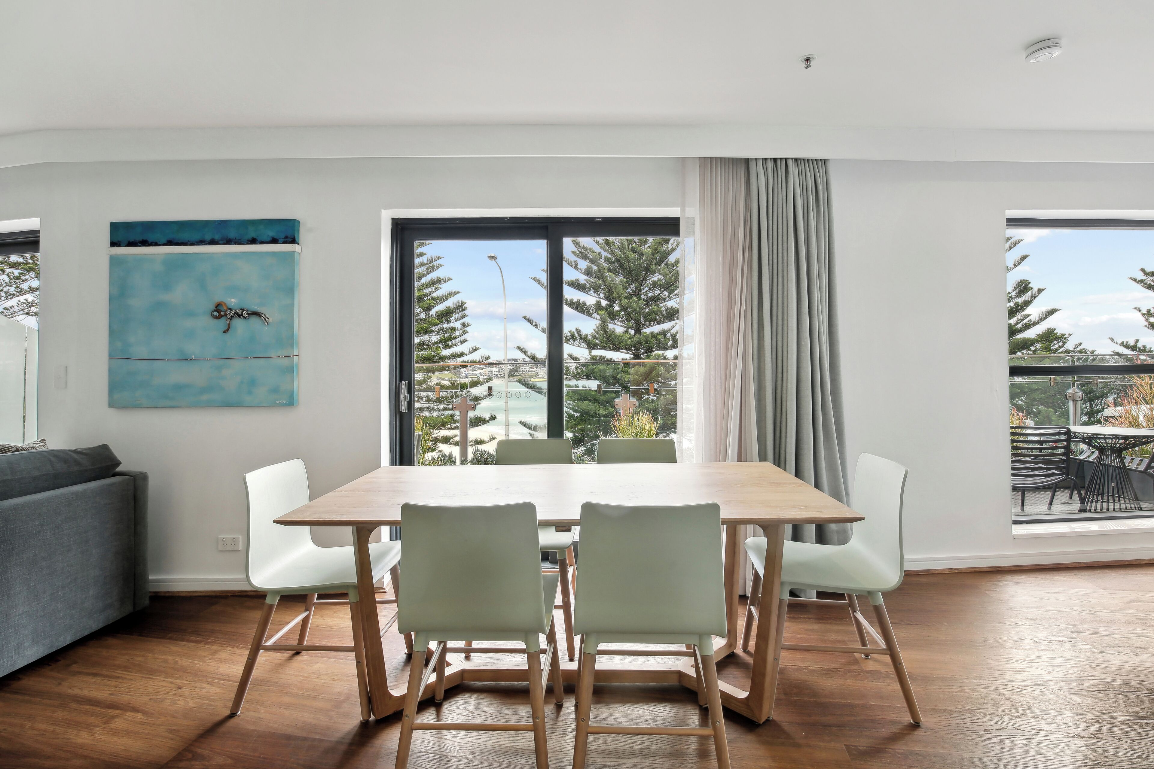 A modern dining area with a wooden table and light-colored chairs, large windows with a view of trees and buildings, and a gray sofa in the background.