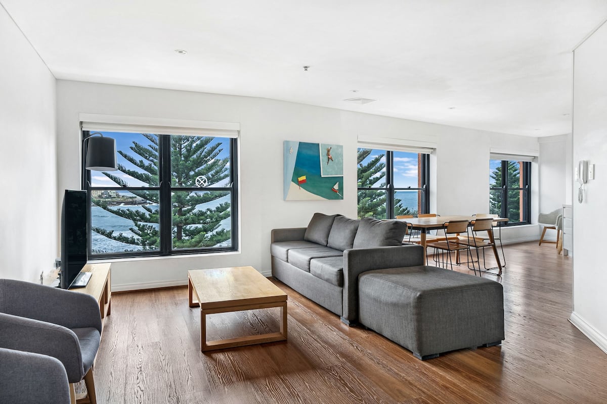 A modern living room with a large window, a grey sofa, a wooden coffee table, and a dining area with chairs.